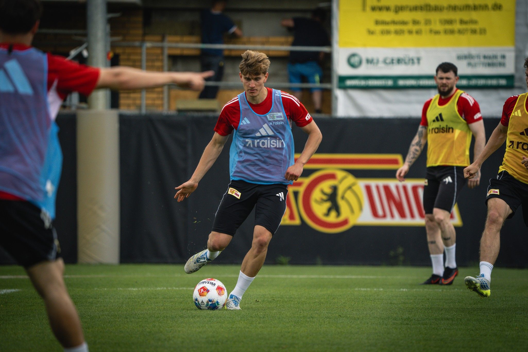 Ein Fußballspieler in Trainingskleidung dribbelt den Ball auf dem Platz, während andere Spieler im Hintergrund üben.
