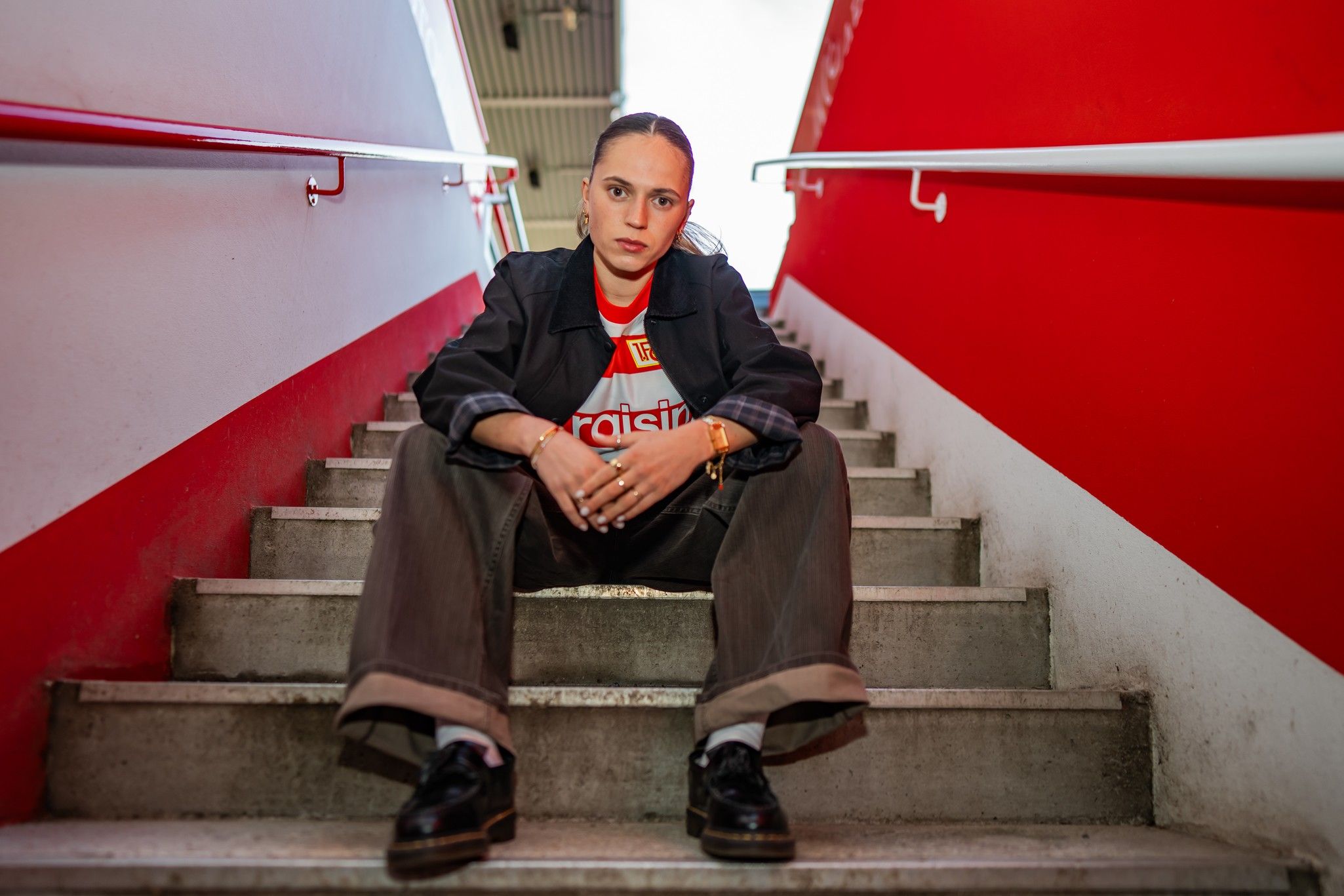 Eine Person sitzt auf einer Treppe, trägt lässige Kleidung und schaut direkt in die Kamera. Rote und weiße Wände umgeben sie.