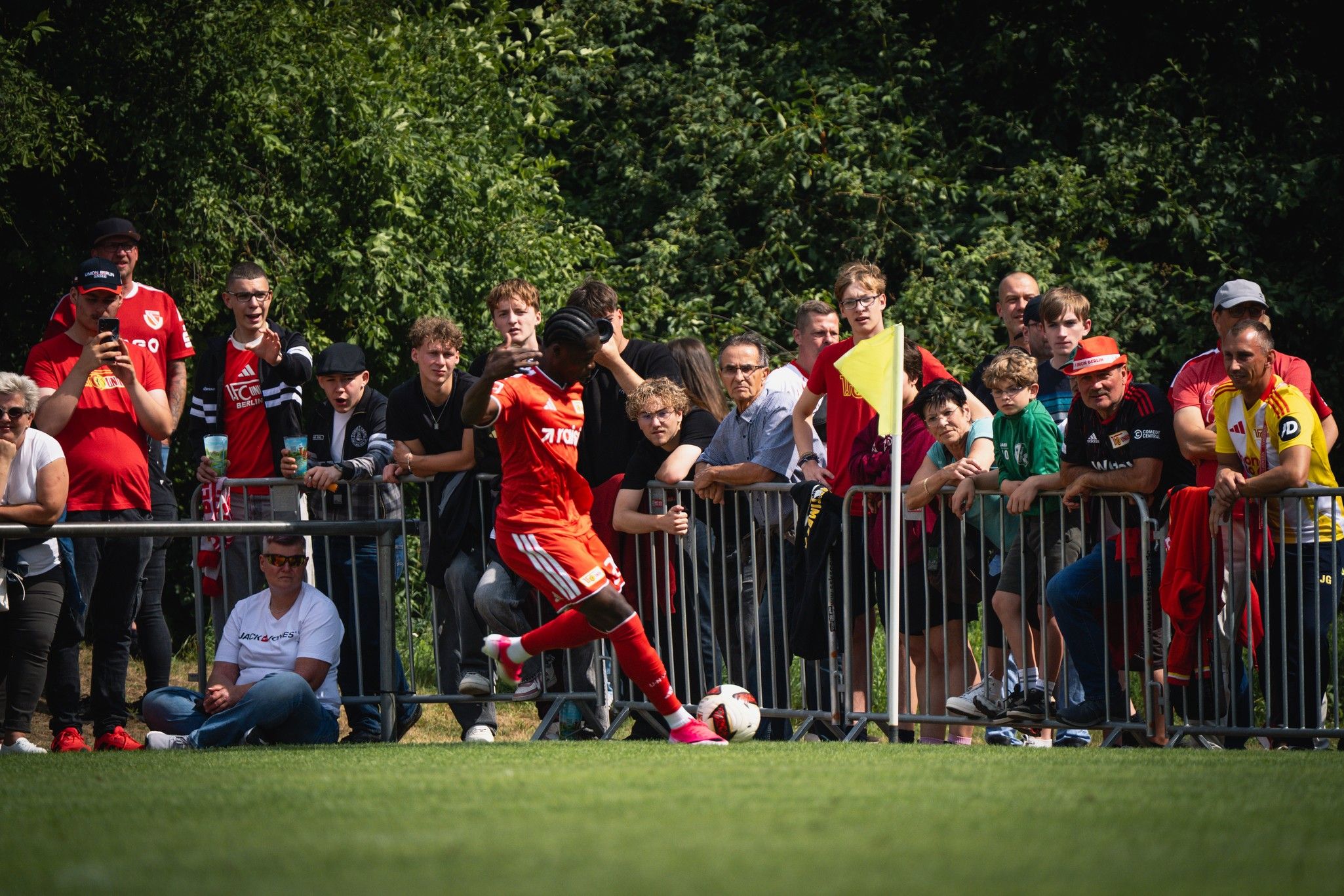 Ein Fußballspieler in roter Kleidung bereitet sich beim Training auf einen Freistoß vor, während Zuschauer zuschauen.