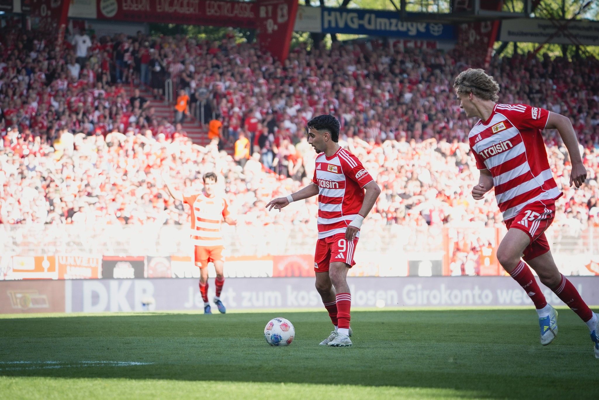 Spielszene eines Fußballspiels mit Spielern in roten Trikots und jubelndem Publikum im Hintergrund.