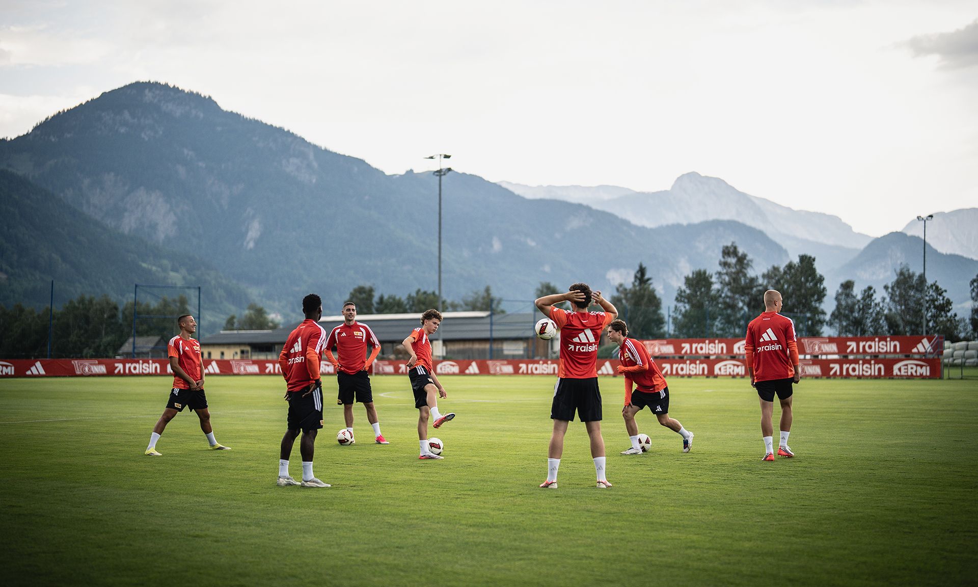 Spieler in roten Trikots trainieren auf einem Fußballfeld mit Bergen im Hintergrund.