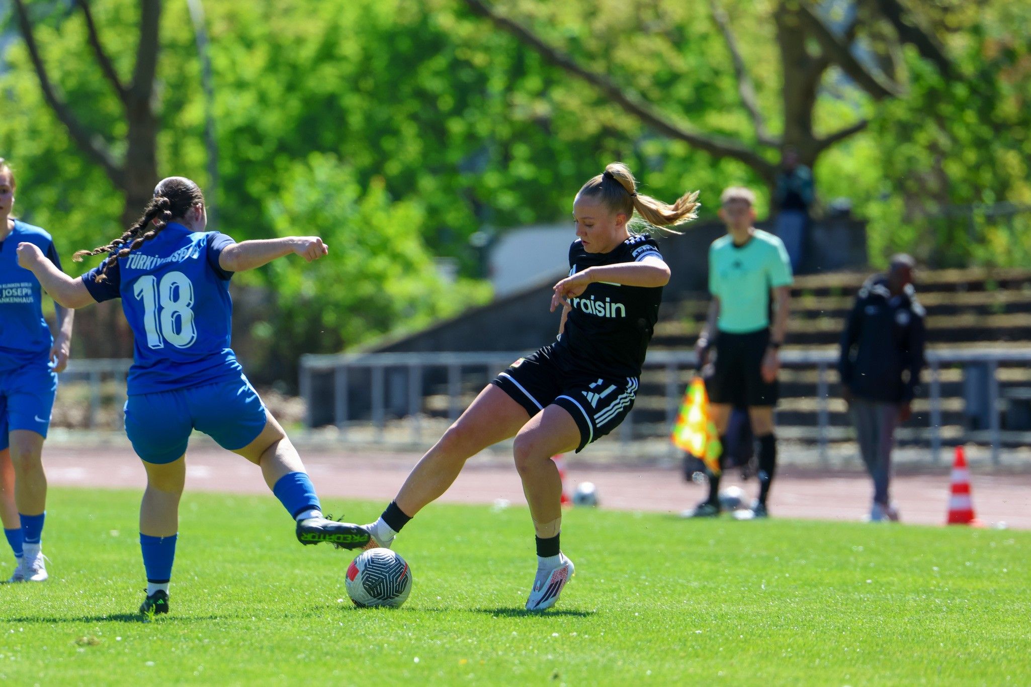 Zwei Frauen im Fußballspiel, eine Spielerin in schwarz tritt nach dem Ball, während die andere in blau attackiert. Hinten Zuschauer und Schiedsrichter.