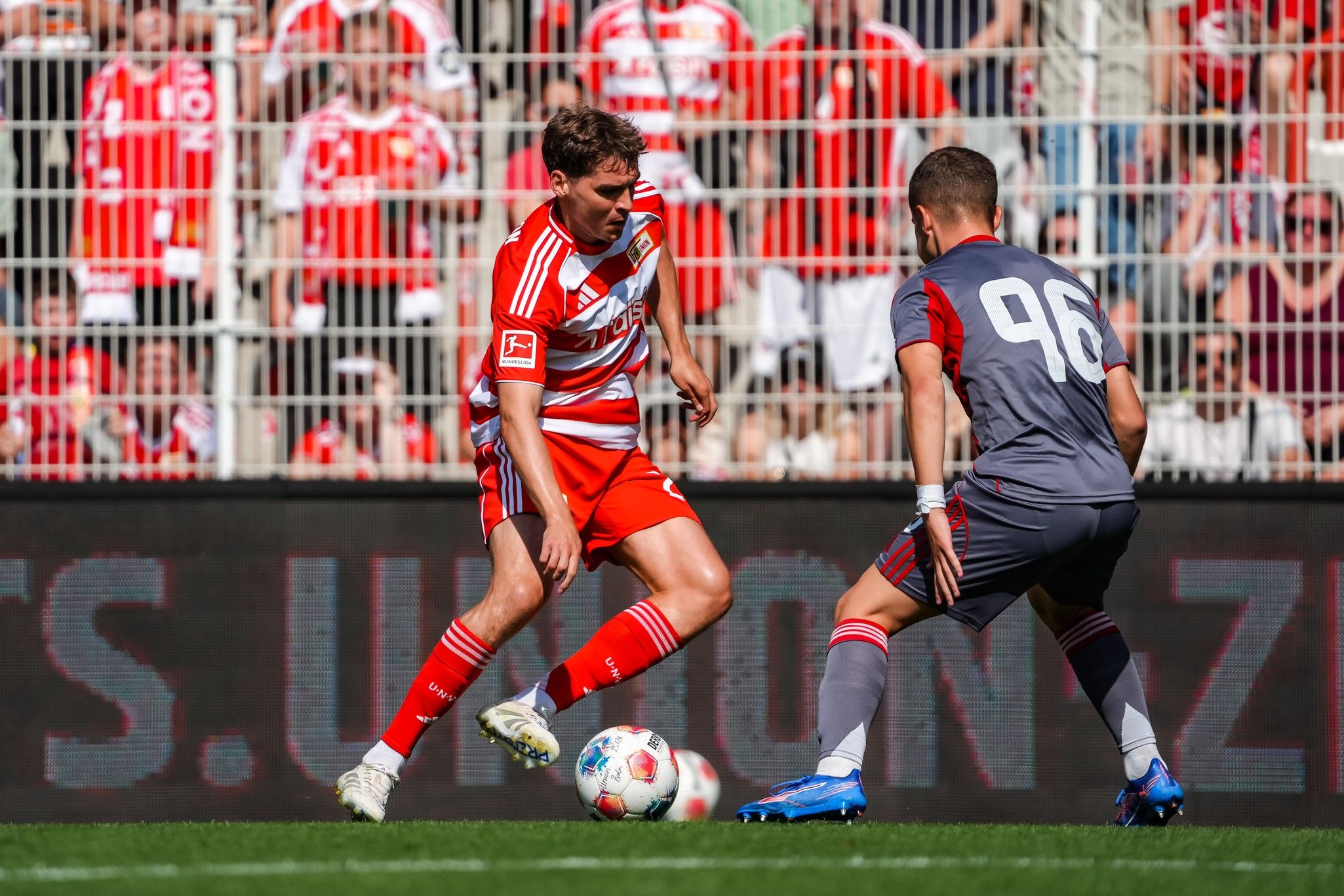 Ein Fußballspiel zeigt einen Spieler in rot-weißer Uniform, der den Ball zu einem Gegner in grauer Uniform dribbelt, im Stadion.