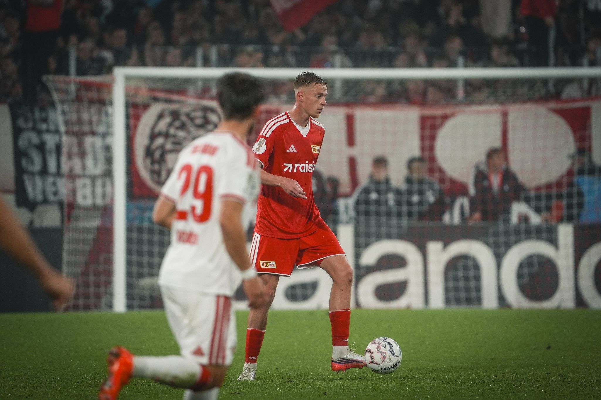 Fußballspieler in rot mit Ball, im Hintergrund Fans und Banner, Stadionatmosphäre.