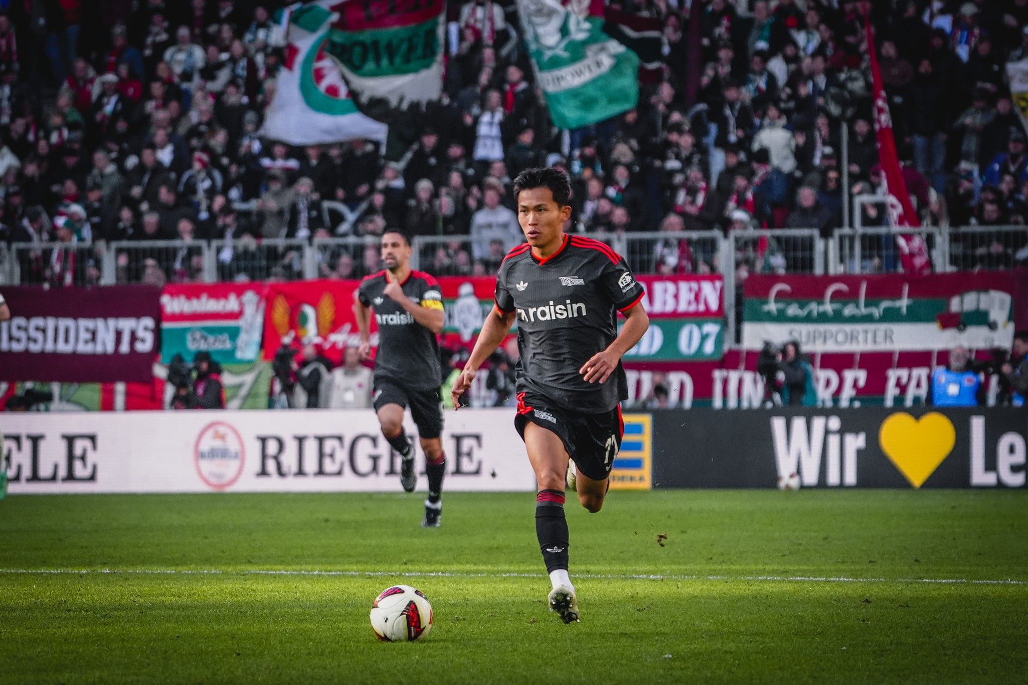 Ein Fußballspieler in grauer Ausrüstung läuft den Ball auf einem Spielfeld entgegen, im Hintergrund jubelnde Fans mit Flaggen.