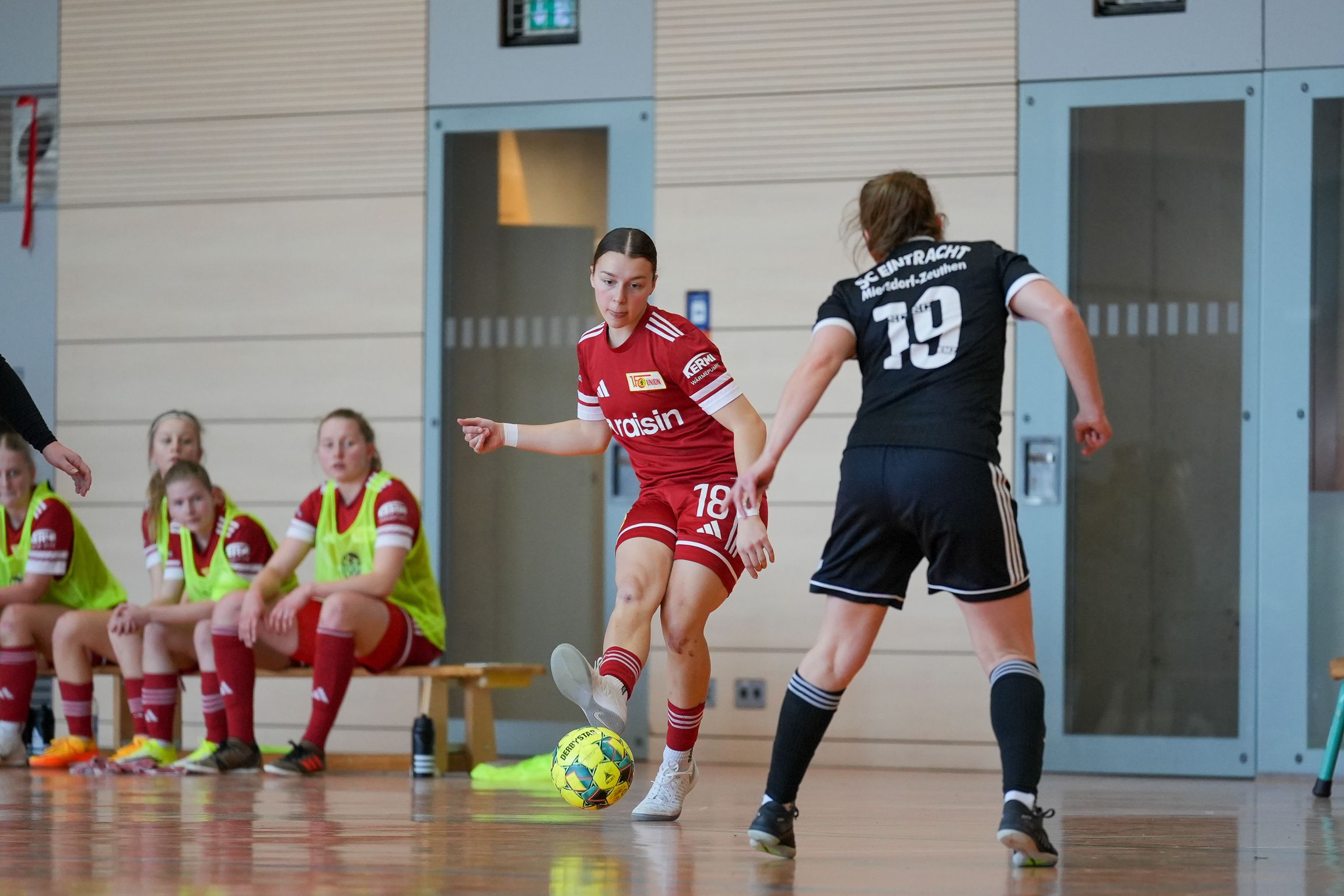 Eine Fußballspielerin in rotem Trikot dribbelt den Ball, während eine Gegnerin in schwarzem Trikot sie verteidigt. Sitzende Spielerinnen im Hintergrund.