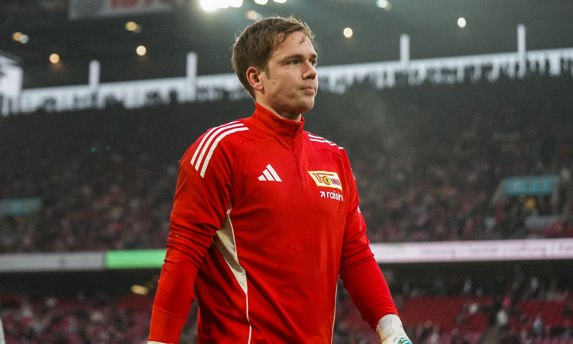 Torwart in rotem Trikot von Union Berlin geht konzentriert auf das Spielfeld, Zuschauer im Hintergrund.