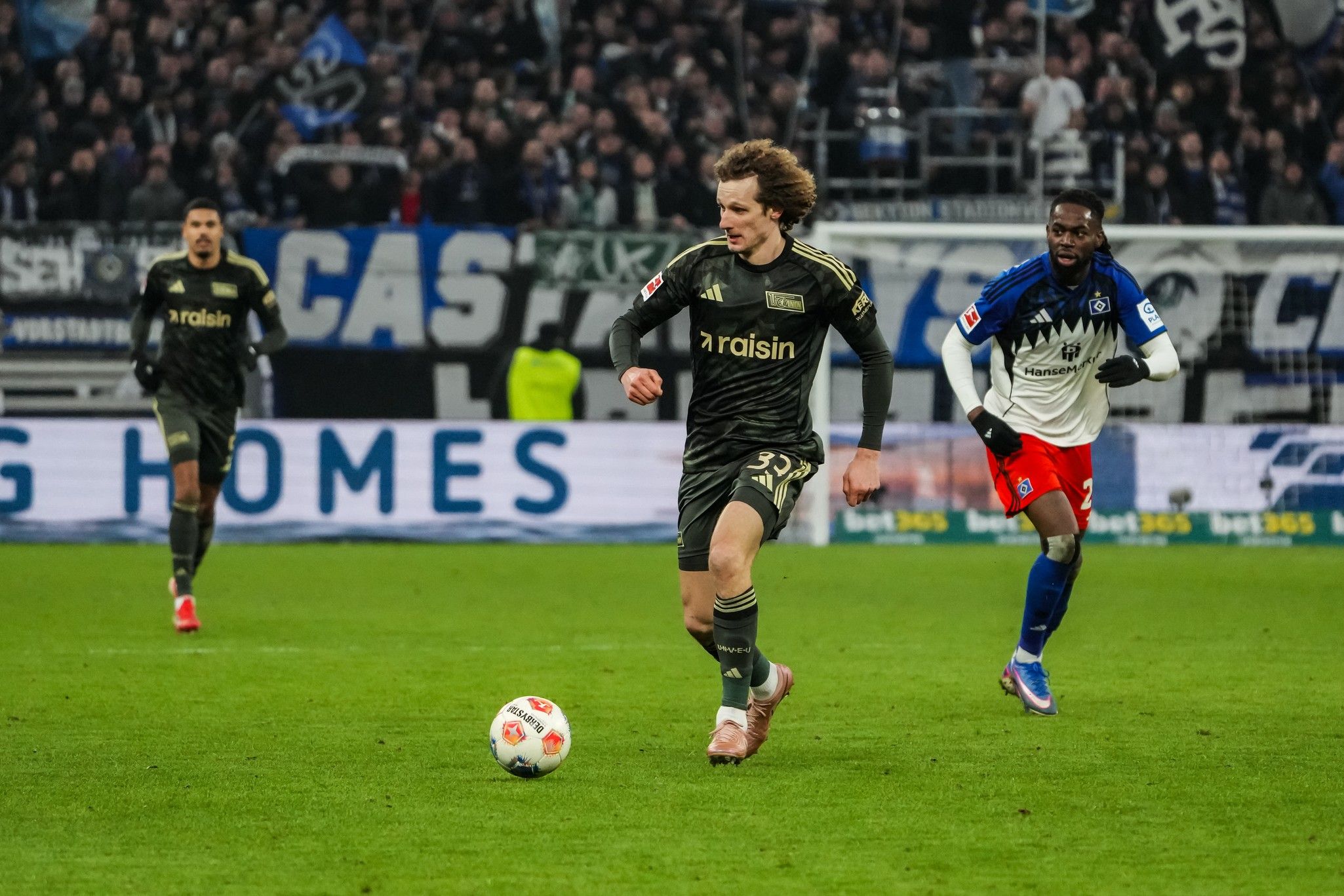 Spielszene aus einem Fußballspiel, in der ein Spieler mit der Nummer 33 einen Ball in Richtung gegnerisches Tor dribbelt, während ein Verteidiger verfolgt.