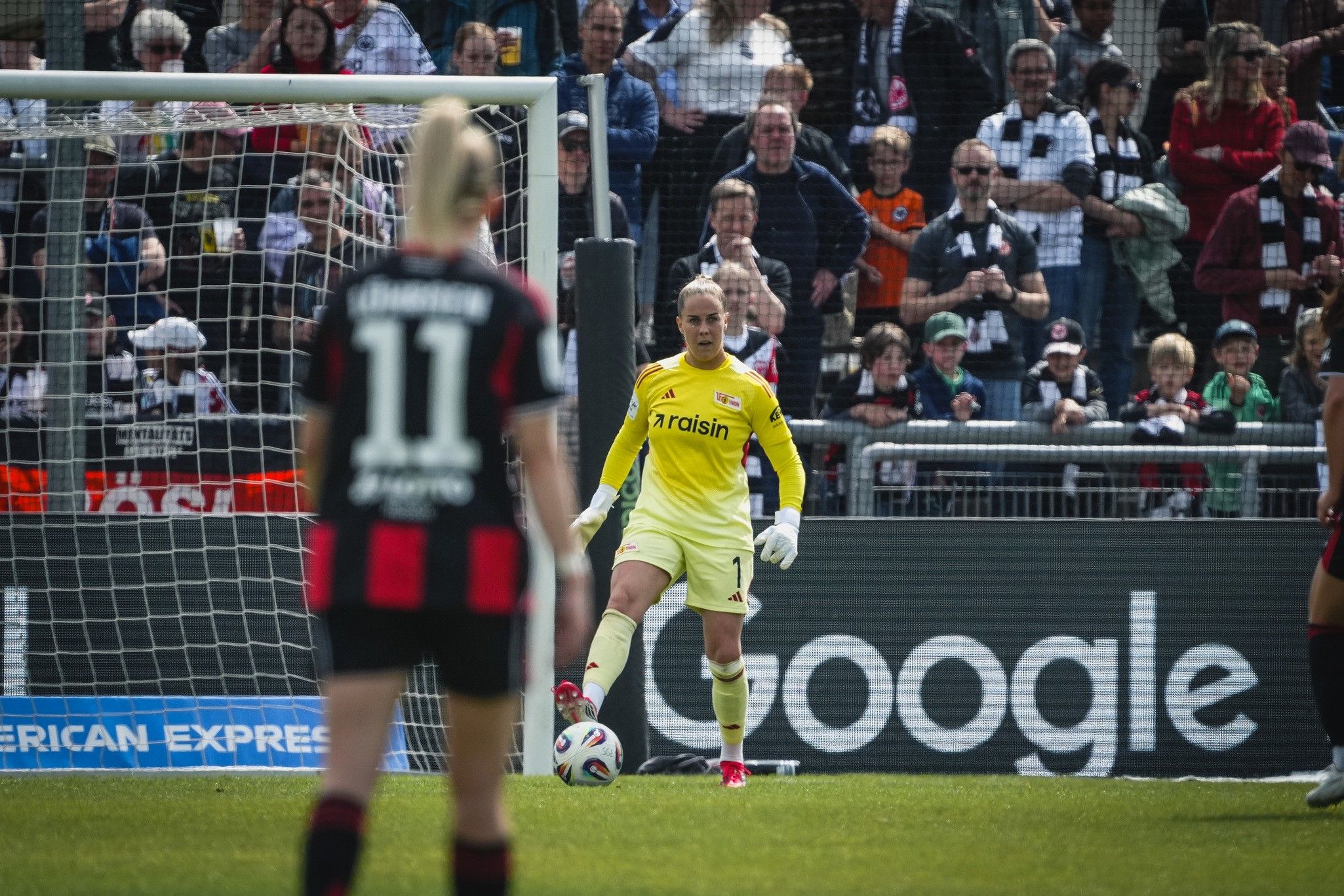 Torfrau in gelber Uniform steht am Tor, Zuschauer im Hintergrund, Spielerin mit der Nummer 11 im Fokus.