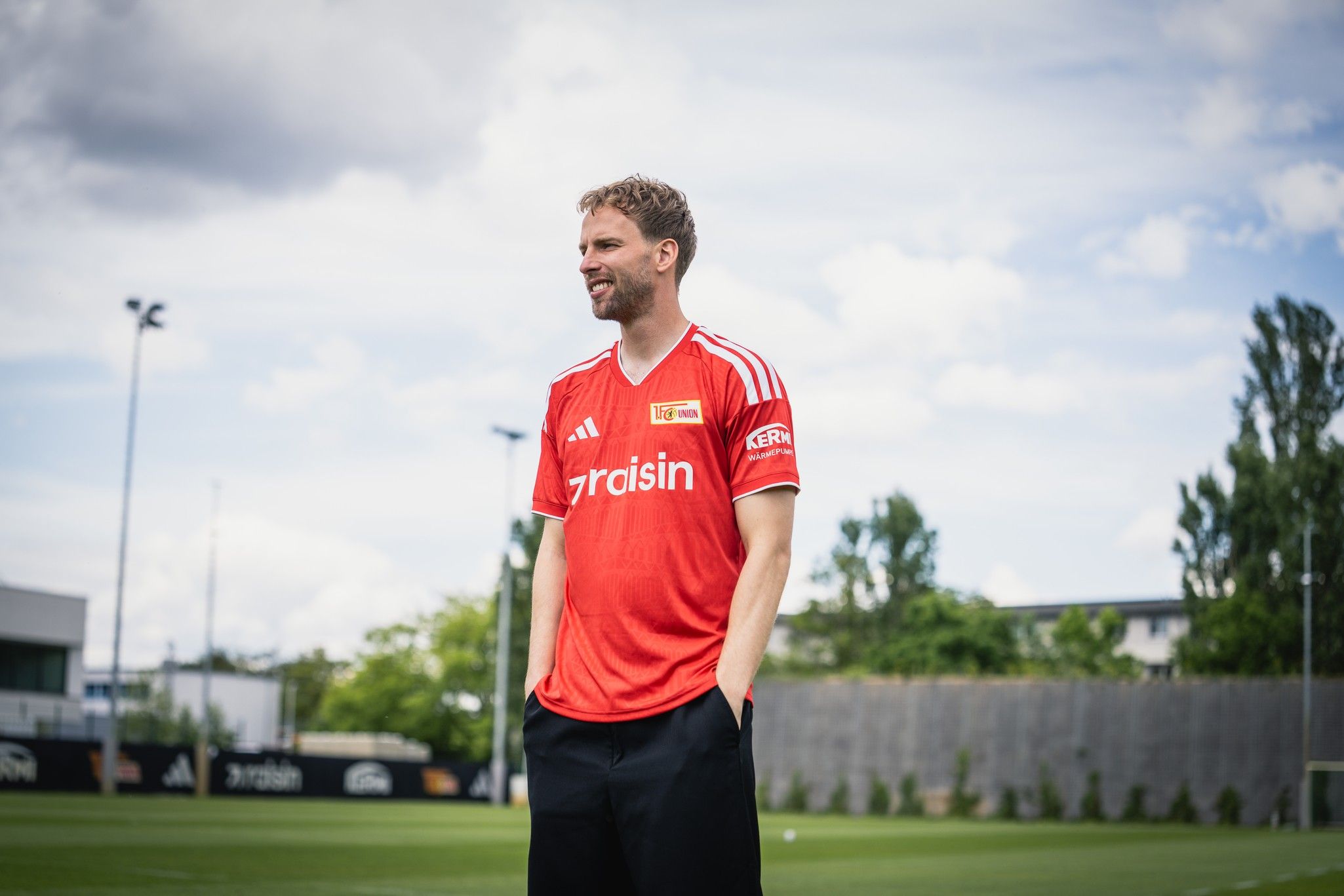 Mann in rotem Fußballtrikot steht auf einem Sportplatz mit grüner Wiese und Bäumen im Hintergrund.