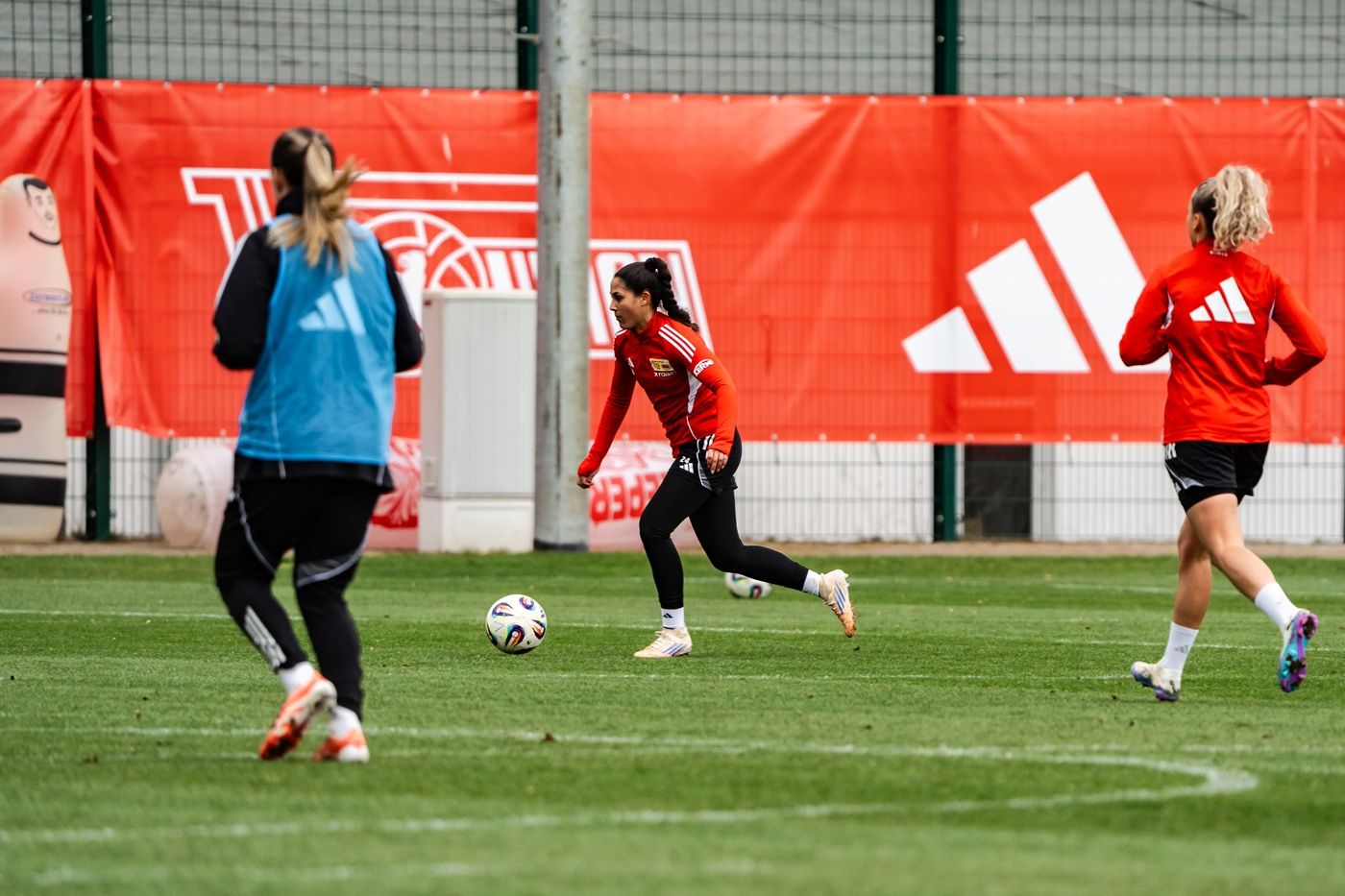 Zwei Fußballspielerinnen üben auf einem Trainingsplatz, während ein rotes Adidas-Banner im Hintergrund sichtbar ist.