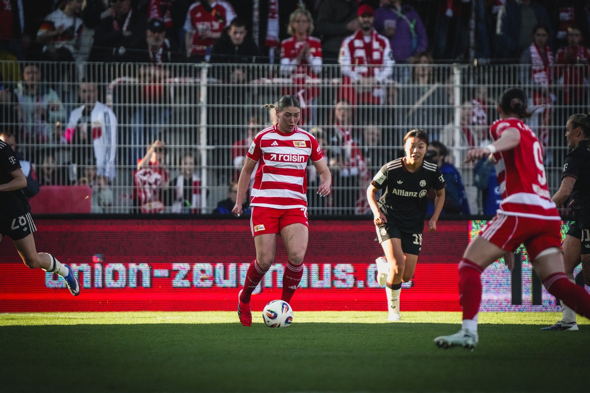 Ein Fußballspieler in rot-weißem Trikot dribbelt den Ball, während andere Spieler in schwarz im Hintergrund stehen. Stadionatmosphäre sichtbar.