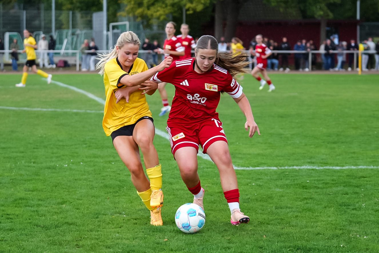 Zwei Fußballspielerinnen in Gelb und Rot kämpfen um den Ball auf einem grünen Spielfeld. Zuschauer stehen im Hintergrund.