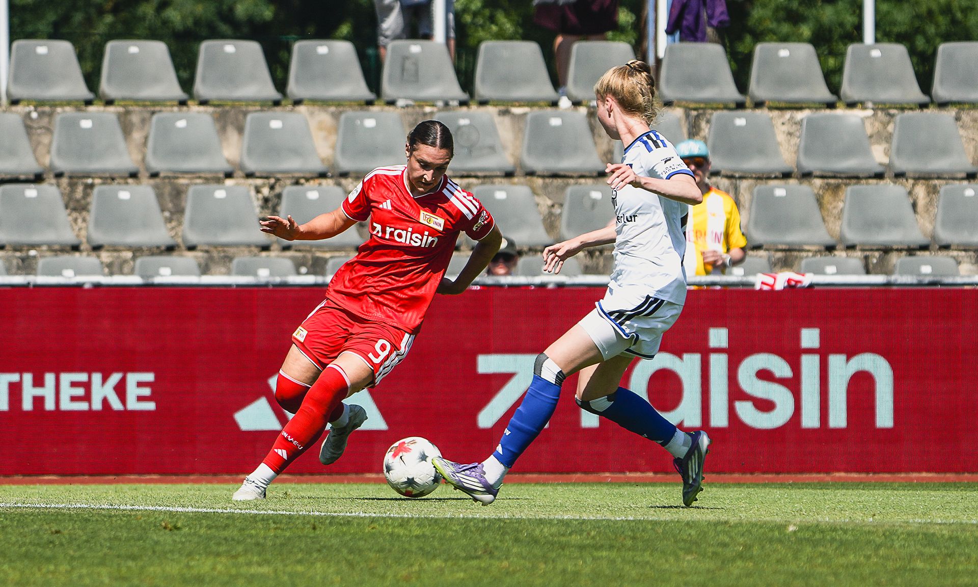 Zwei Fußballspielerinnen im Wettkampf auf dem Spielfeld, eine in rotem Trikot, die andere in weißem Trikot.