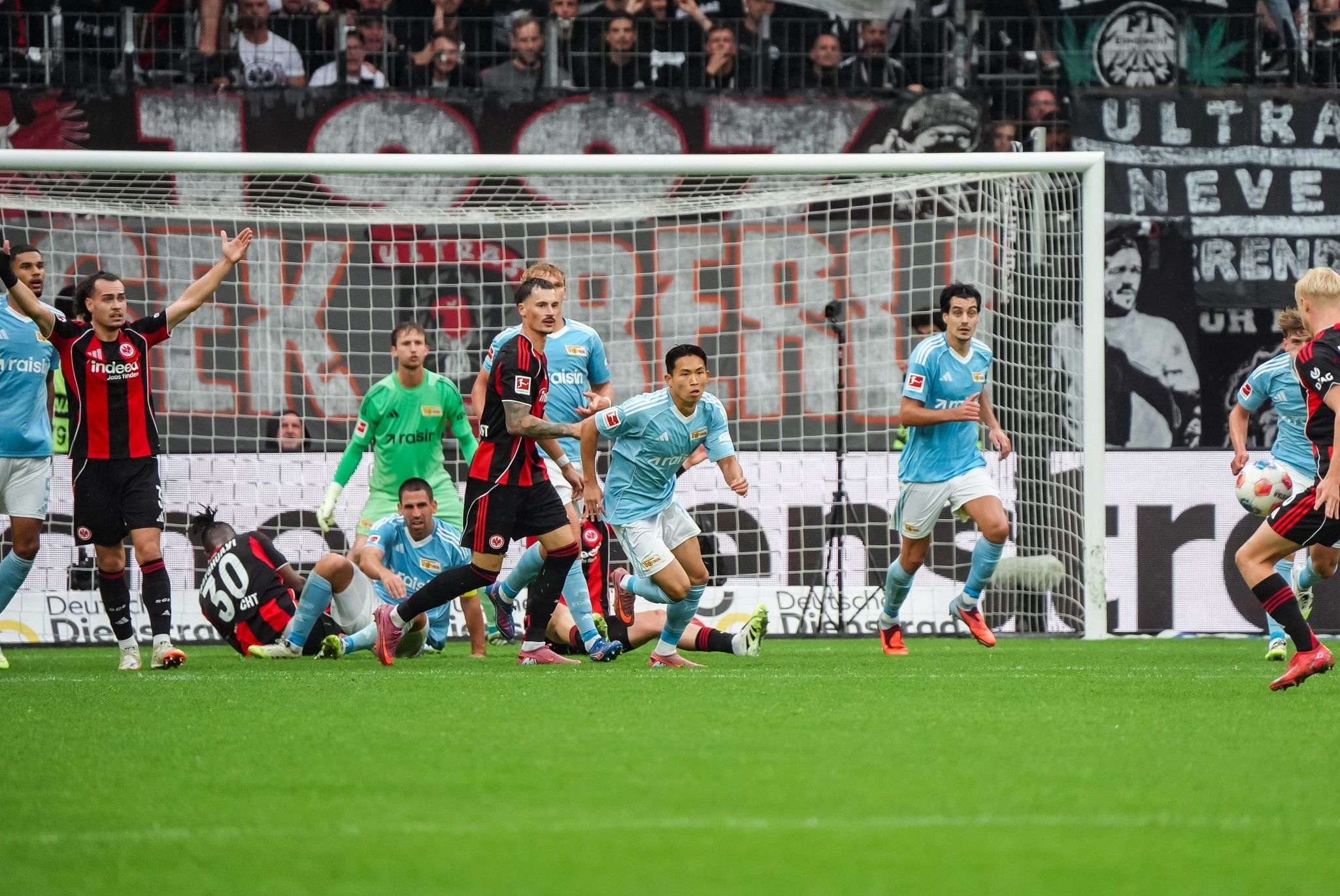Fußballspiel mit Spielern in roten und blauen Trikots, die auf ein Tor zusteuern, im Hintergrund sind Fans und Banner zu sehen.