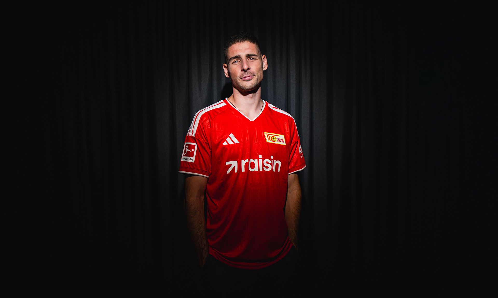 Ein junger Mann trägt ein rotes Fußballtrikot mit dem Logo eines Vereins und steht vor einem dunklen Hintergrund.