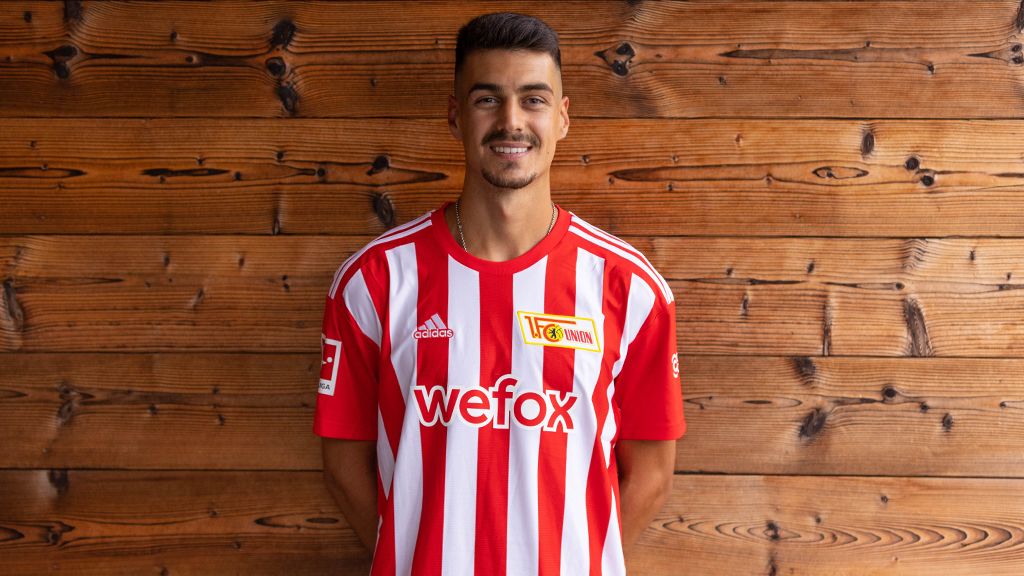 Ein junger Fußballspieler trägt ein rot-weiß gestreiftes Trikot mit Sponsorenlogo vor einer Holzwand.