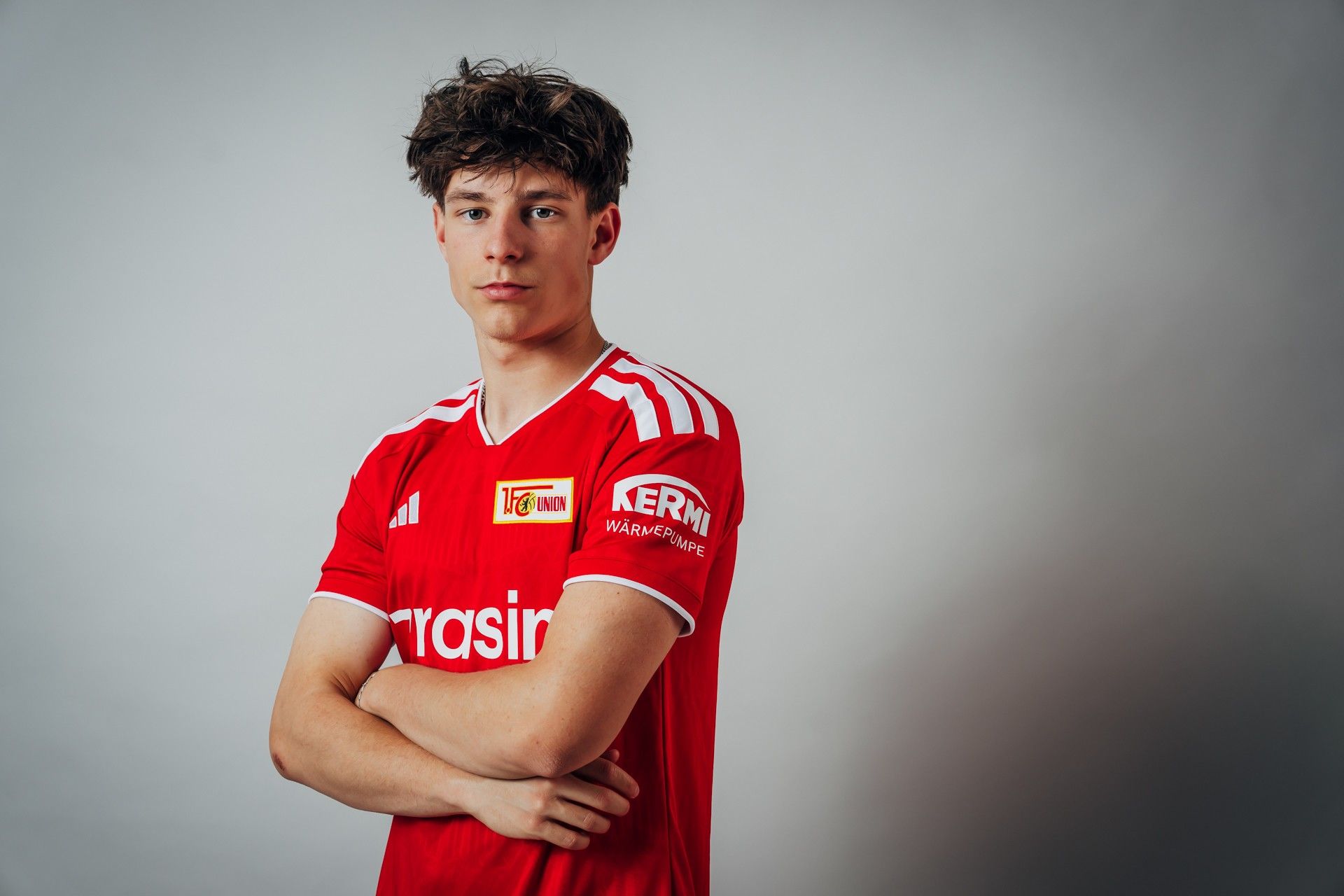 Ein junger Mann mit gewelltem Haar steht in einem roten Fußballtrikot mit einem Sponsorenschriftzug und verschränkten Armen vor einem neutralen Hintergrund.
