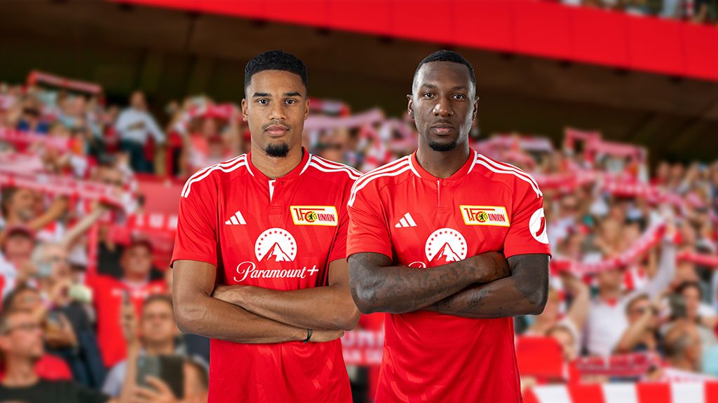Zwei Fußballspieler in roten Trikots stehen mit verschränkten Armen vor einer jubelnden Menschenmenge im Stadion.
