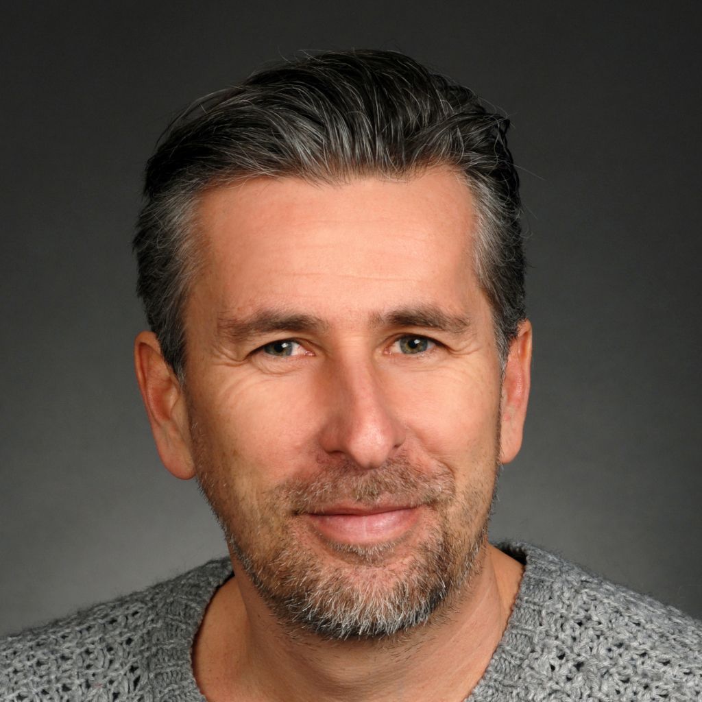 Ein Mann mit grauen Haaren und Bart, der in einem grauen, grob gestrickten Pullover lächelt.