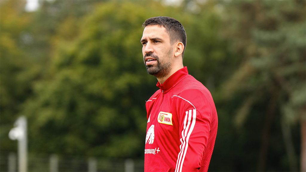 A football player in a red tracksuit stands outdoors, looking thoughtfully into the distance, surrounded by green trees.