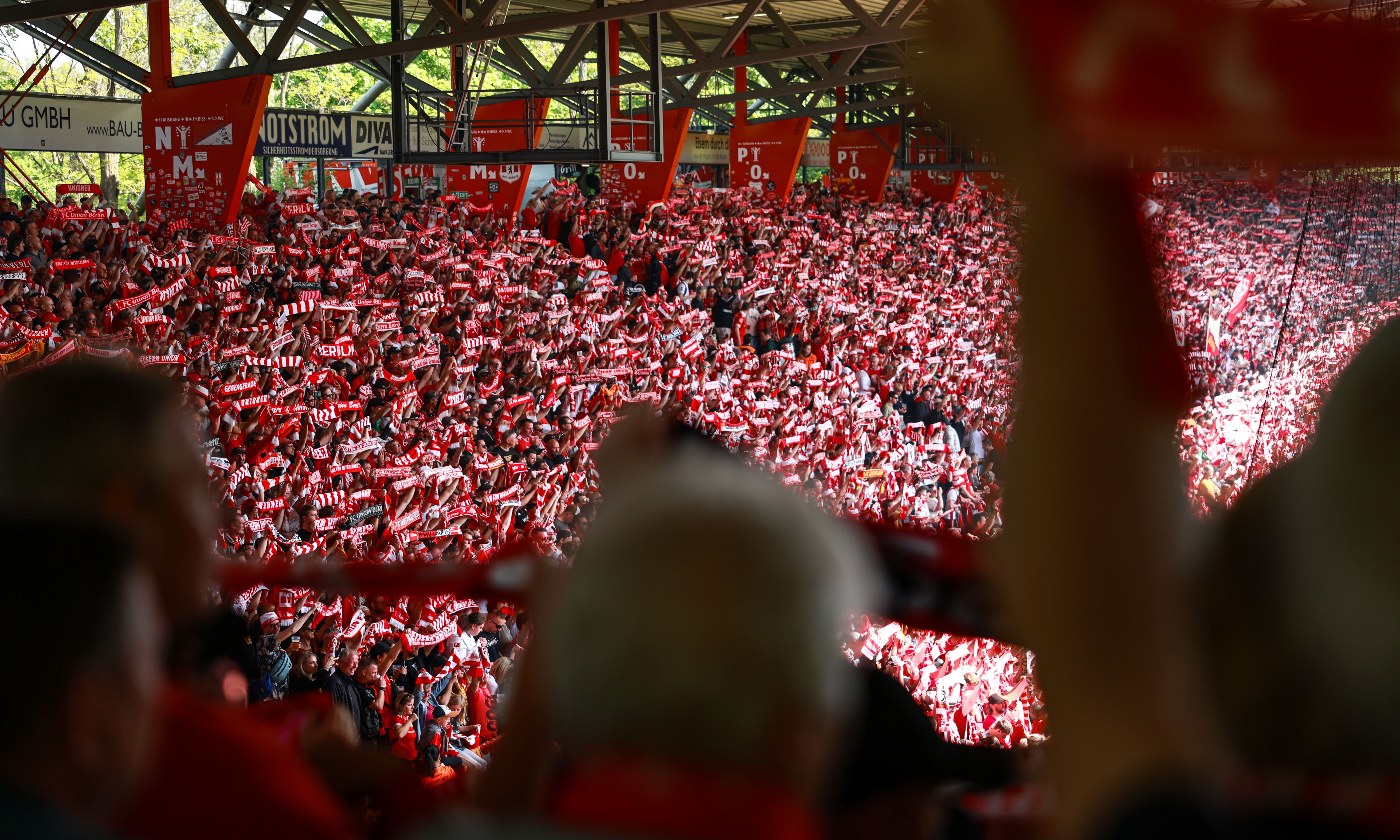 Ein Stadion voller jubelnder Fans in roten Trikots, die Schals in der Luft halten.