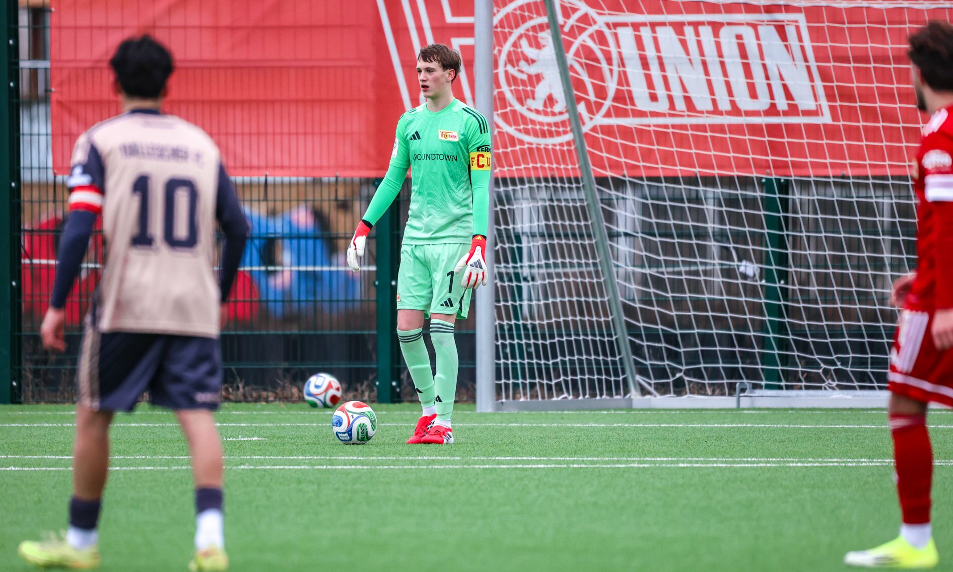 Torwart in grüner Ausrüstung steht vor dem Tor auf einem Fußballfeld, während Spieler im Hintergrund agieren.