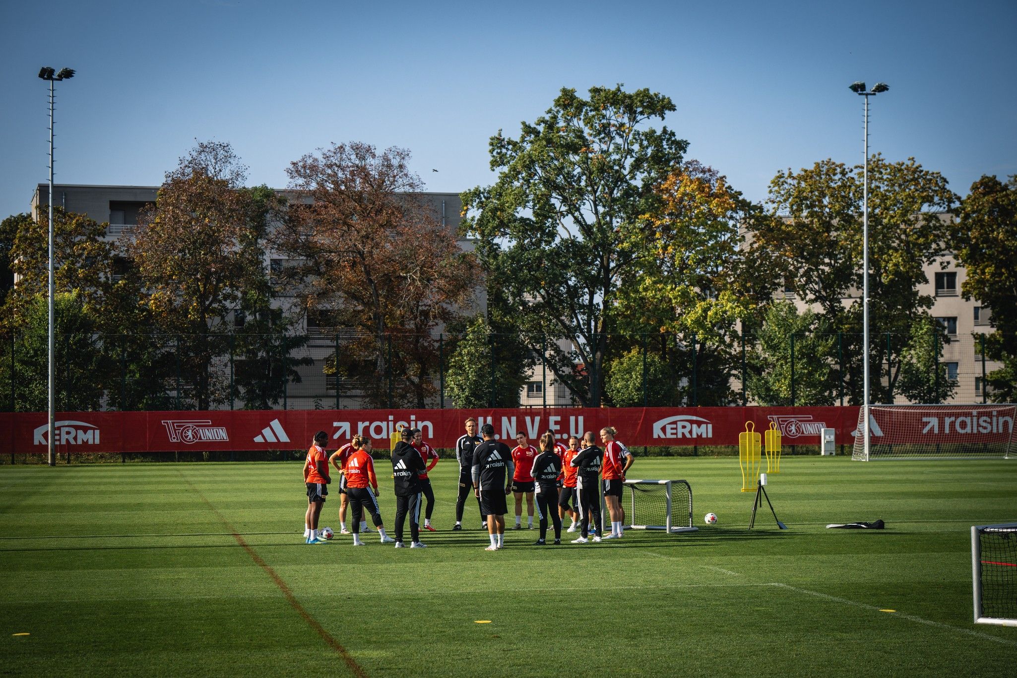 Eine Gruppe von Personen in Sportkleidung trainiert auf einem Fußballplatz mit Toren und Bäumen im Hintergrund.