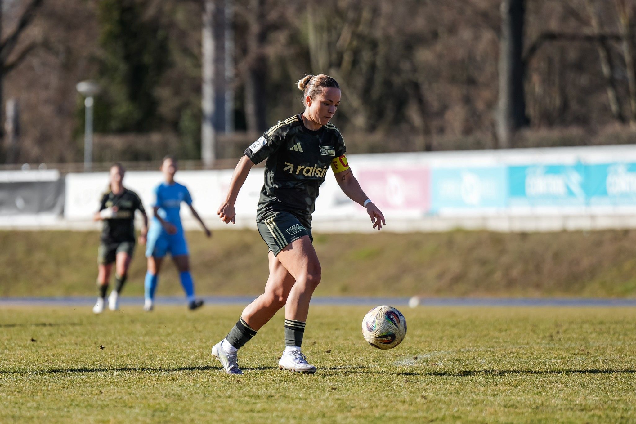 Eine Fußballspielerin schießt einen Ball auf einem Rasenplatz, während andere Spielerinnen im Hintergrund stehen.