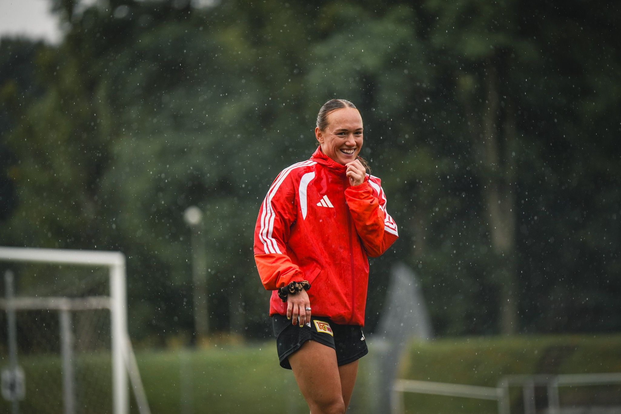 Eine sportlich gekleidete Frau mit Kurzhaarschnitt steht lächelnd im Regen auf einem Fußballplatz.