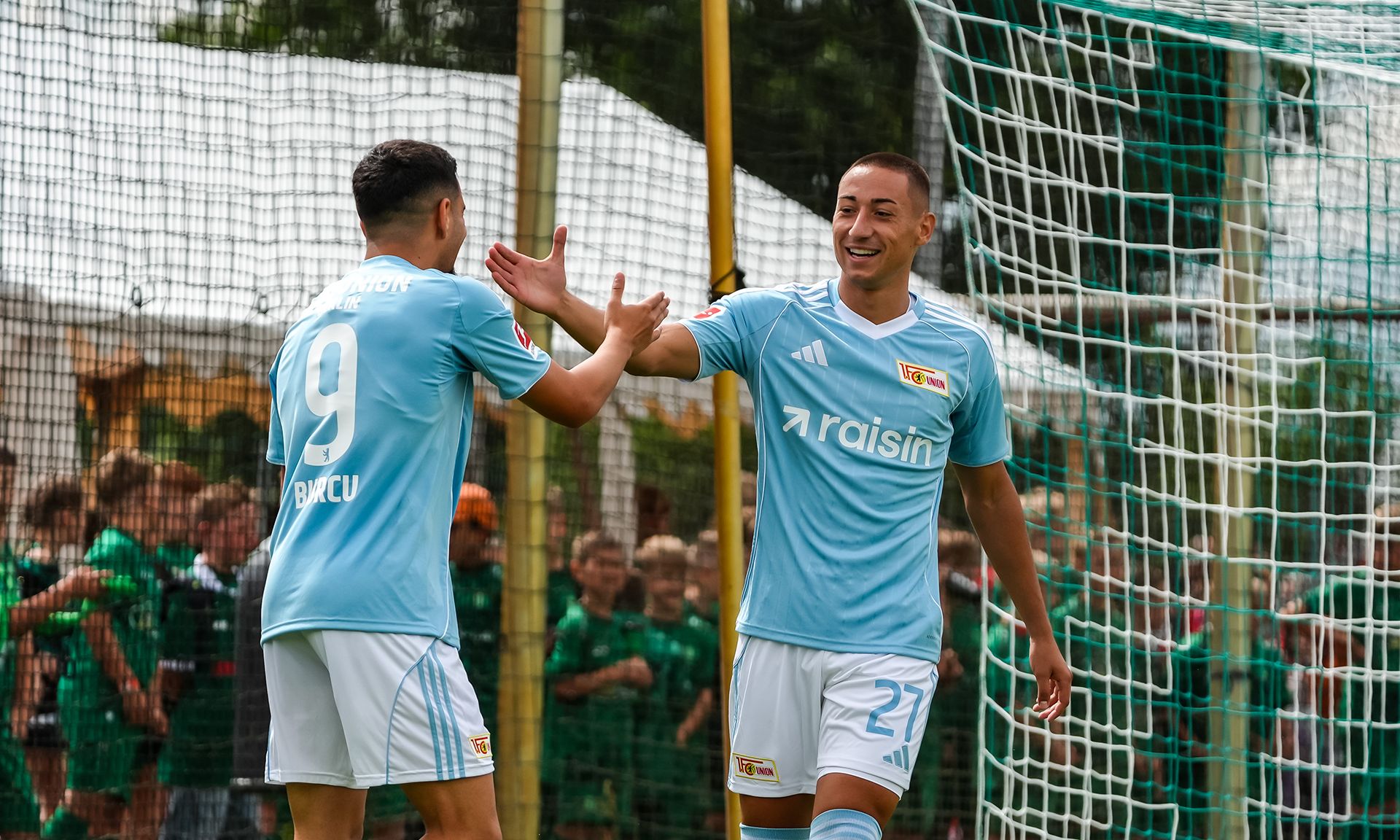 Zwei Fußballspieler in hellblauen Trikots feiern einen Torerfolg vor einem Fußballtor.