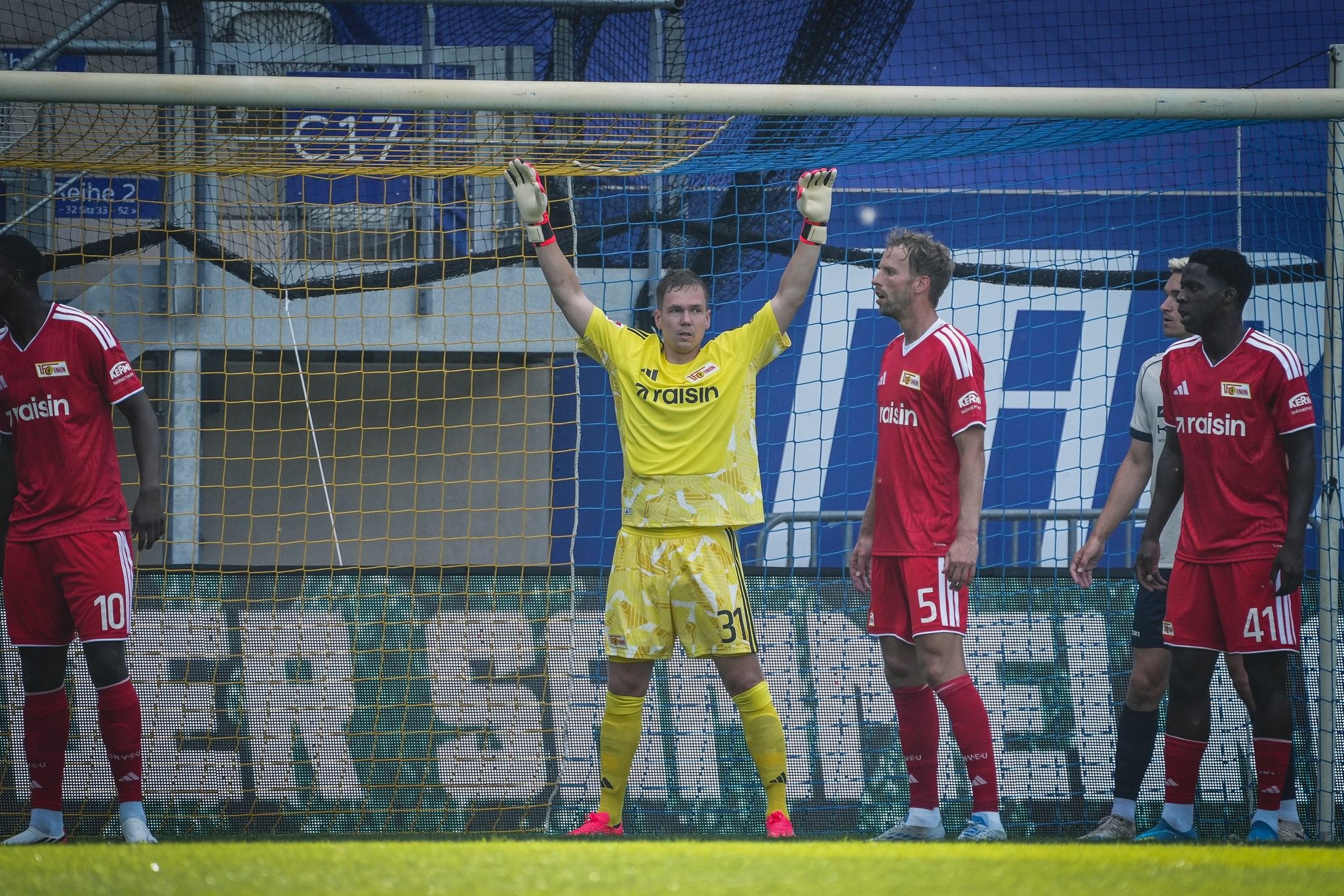 Der Torwart steht bereit im Tor, während seine Mitspieler in roten Trikots das Spielfeld im Hintergrund beobachten.