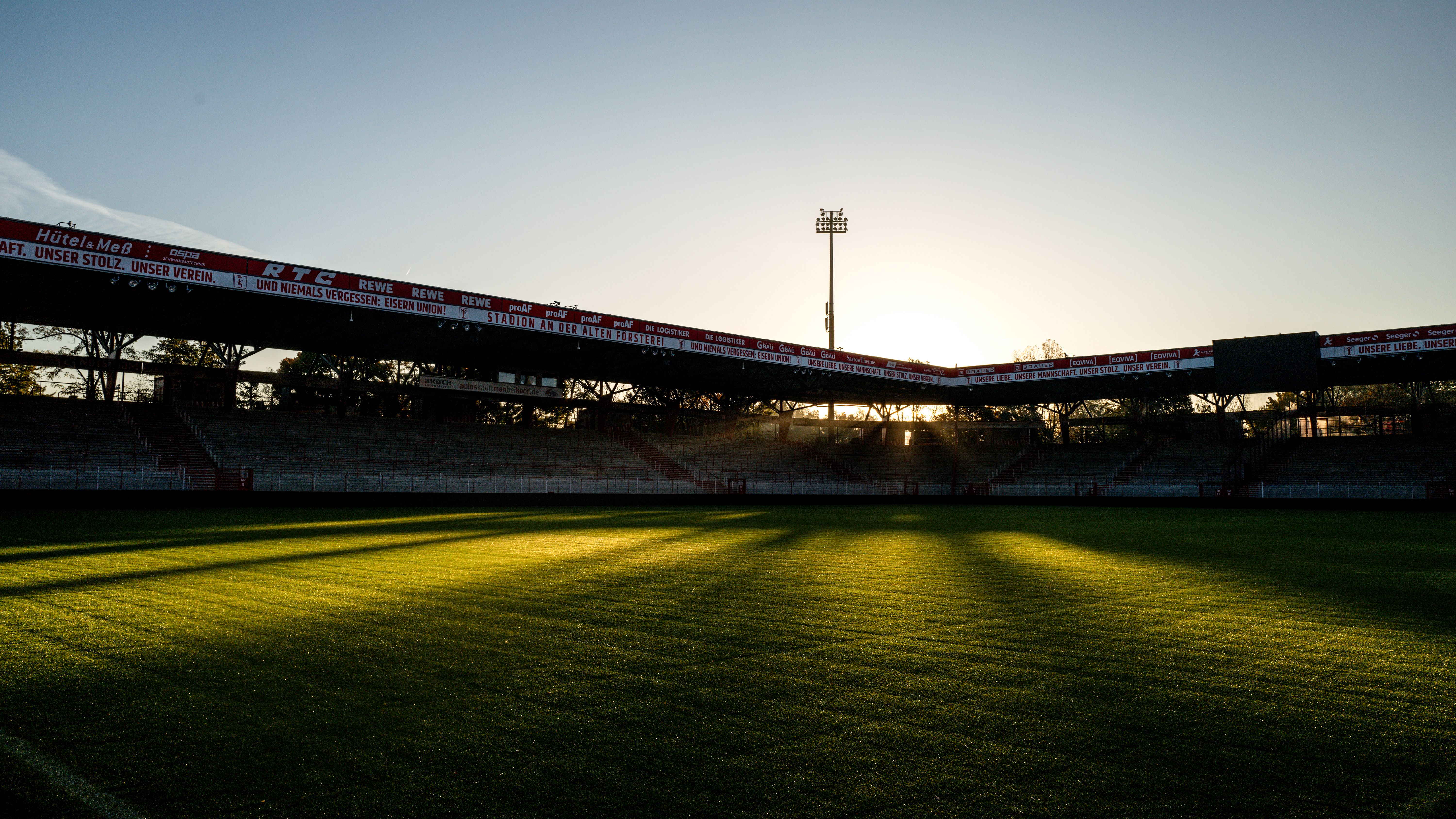 Stadion mit leuchtendem Grasfeld und Sonnenuntergang im Hintergrund, dunkle Schatten entlang der Tribünen.