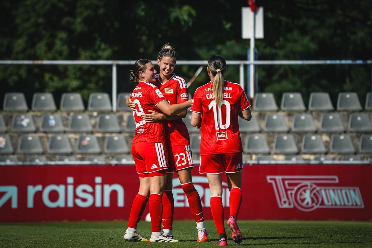 Drei Spielerinnen in roten Trikots umarmen sich jubelnd auf einem Fußballfeld. Im Hintergrund sind leere Zuschauerplätze zu sehen.