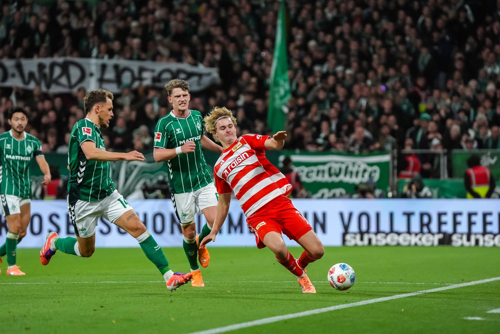 Ein Fußballspieler fällt während eines Spiels, während zwei Gegner in grün hinter ihm stehen. Stadionatmosphäre mit Fans im Hintergrund.