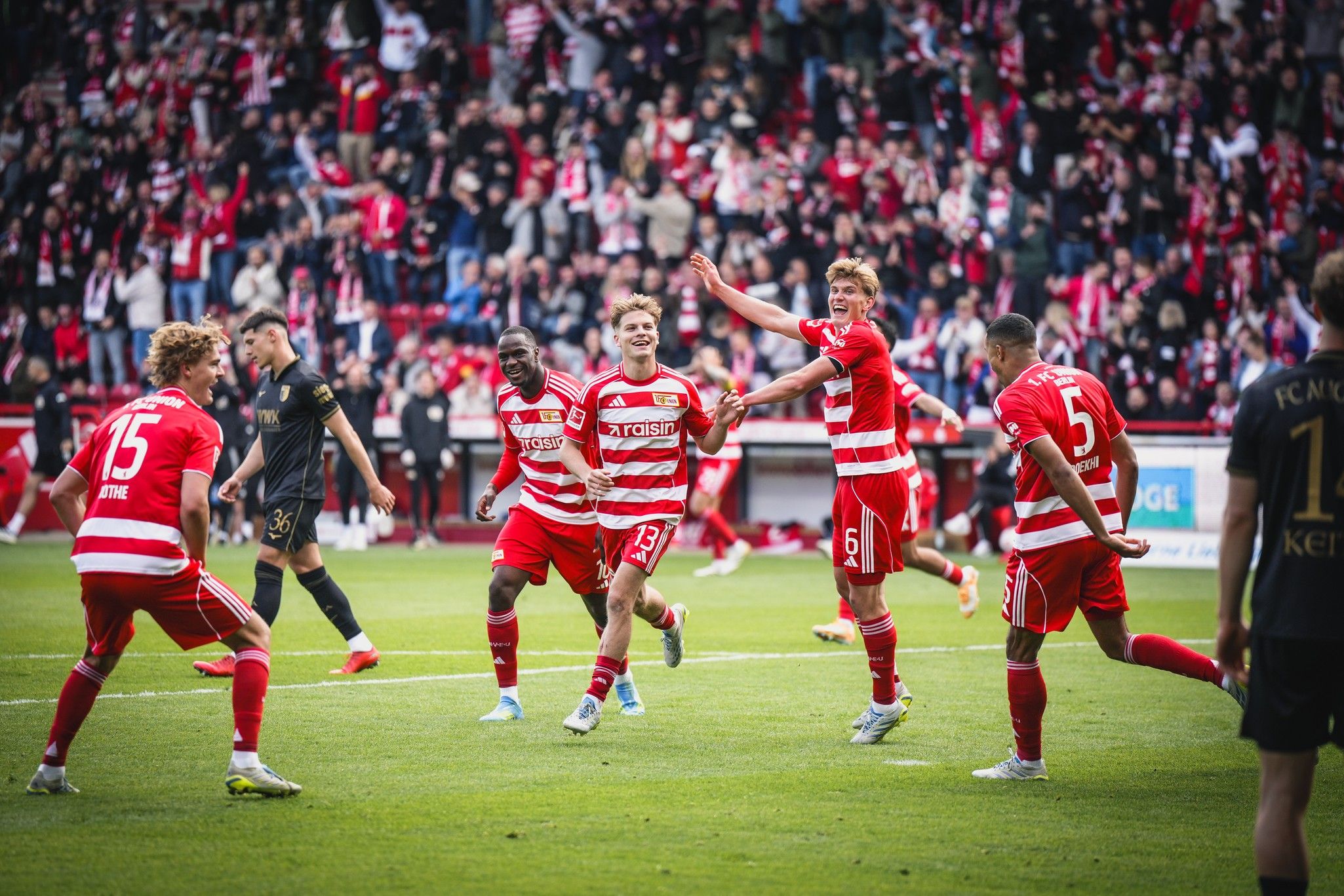 Jubiliate Fußballspieler in rot-weiß gestreiften Trikots feiern ein Tor vor begeisterten Fans im Hintergrund.