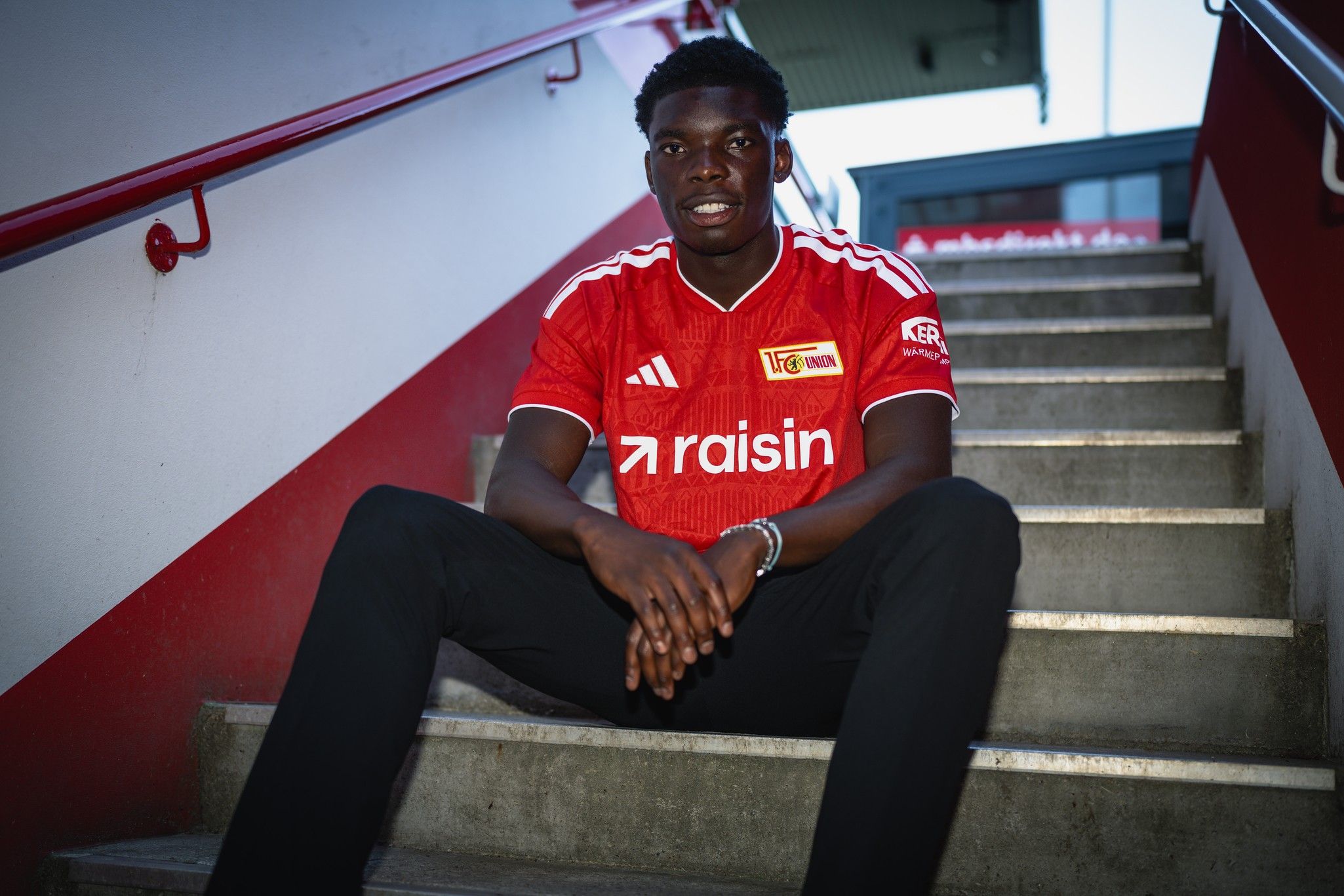 Ein junger Mann sitzt auf einer Treppe und trägt ein rotes Fußballtrikot mit dem Schriftzug „raisin“.