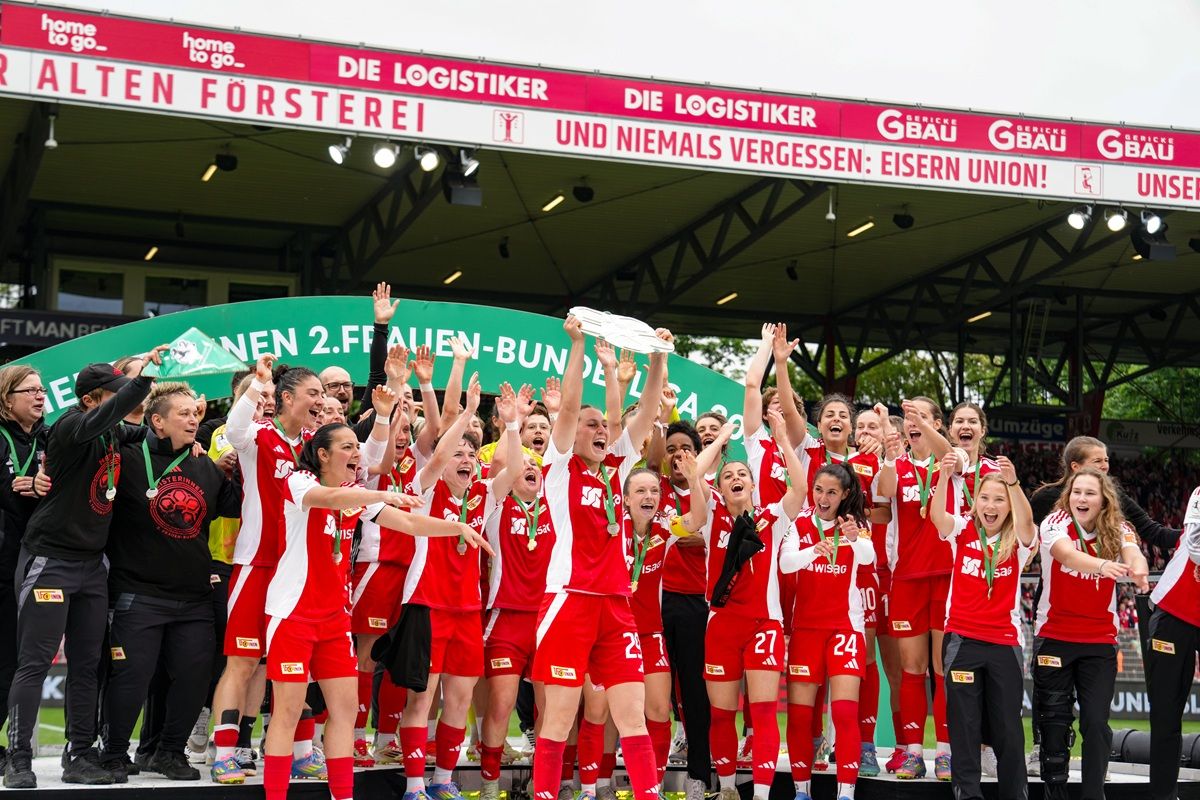 Das Bild zeigt ein jubelndes Fußballteam mit Pokal, das auf einem Siegertreppchen steht. Fans applaudieren im Hintergrund.