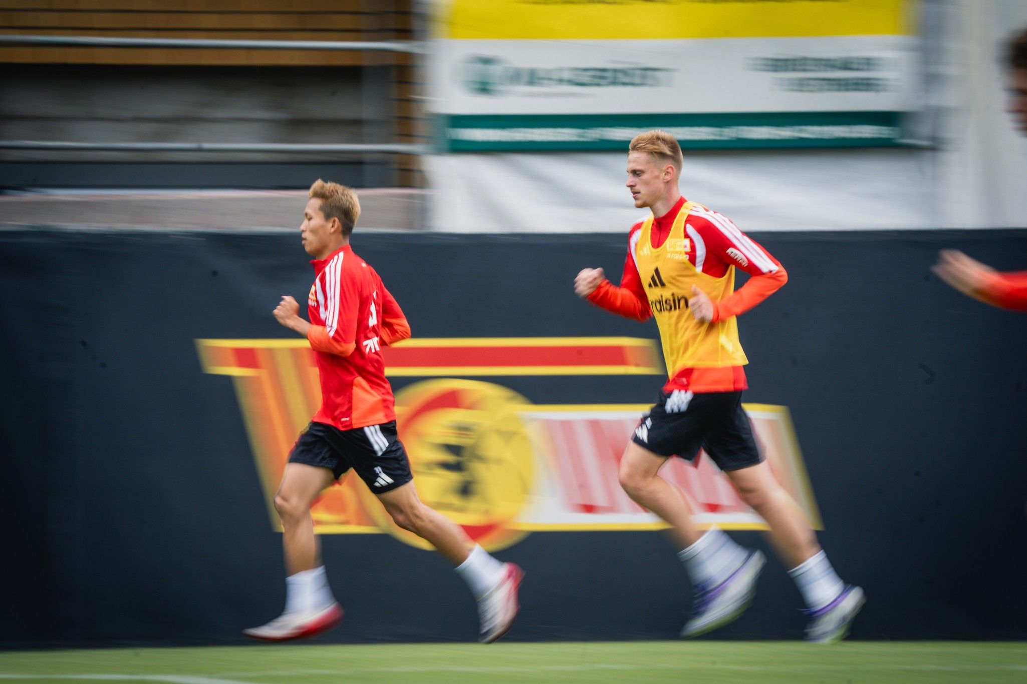 Zwei Fußballspieler joggen im Training auf einem Platz, während ein dritter Spieler im Hintergrund läuft.