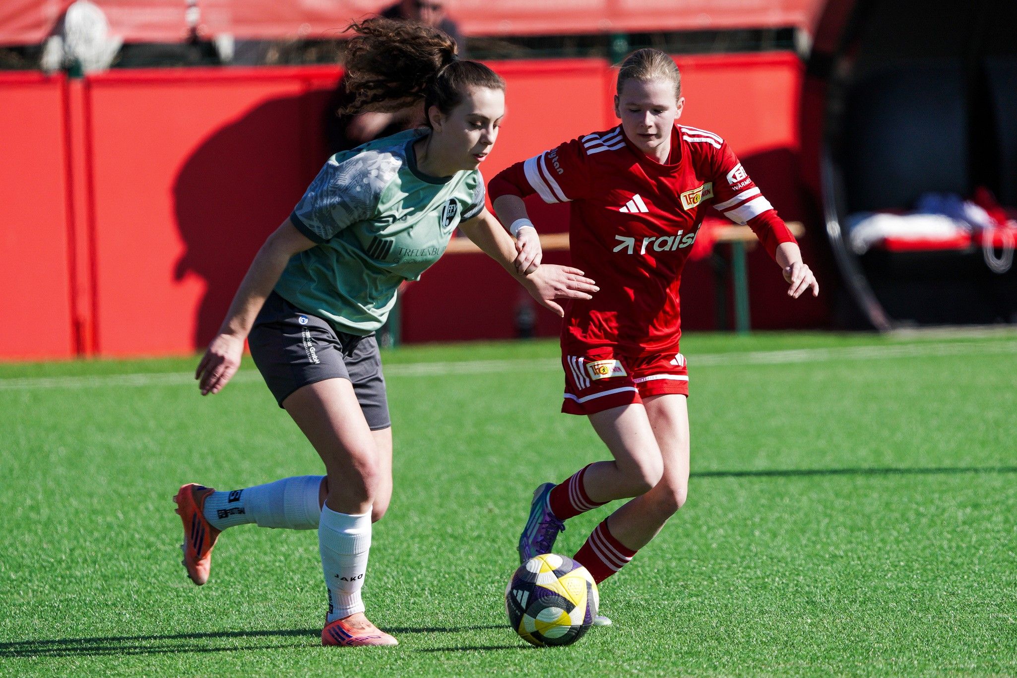 Zwei Frauen im Fußballspiel sprinten dem Ball hinterher, eine trägt grüne und die andere rote Trikots auf einem Kunstrasenplatz.
