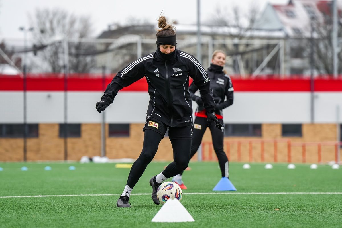 Eine Sportlerin in Trainingskleidung dribbelt einen Fußball auf einem Kunstrasenplatz. Im Hintergrund ist eine andere Spielerin zu sehen.