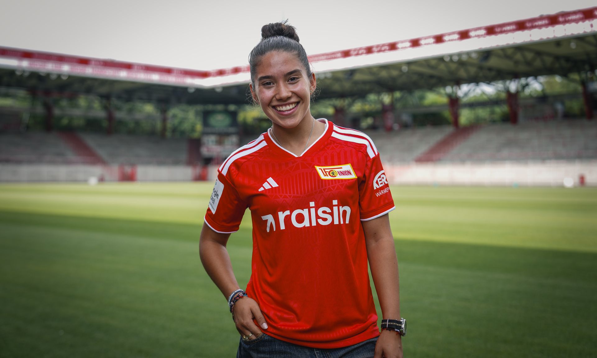 Eine lächelnde Frau trägt ein rotes Fußballtrikot mit dem Logo von Union Berlin und steht auf einem grünen Fußballfeld.