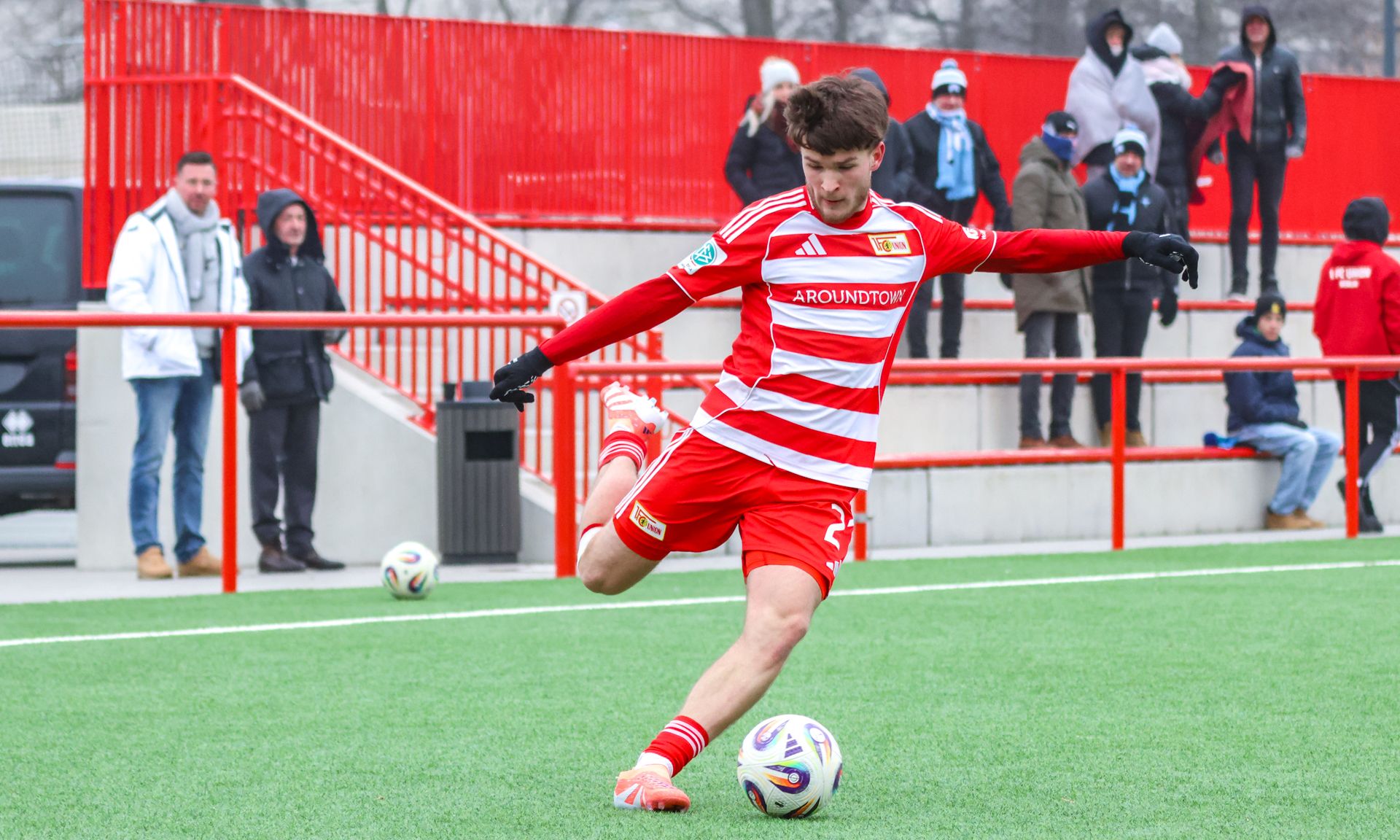 Ein junger Fußballspieler in rotem Trikot schießt den Ball auf einem Kunstrasenplatz. Zuschauer stehen im Hintergrund.