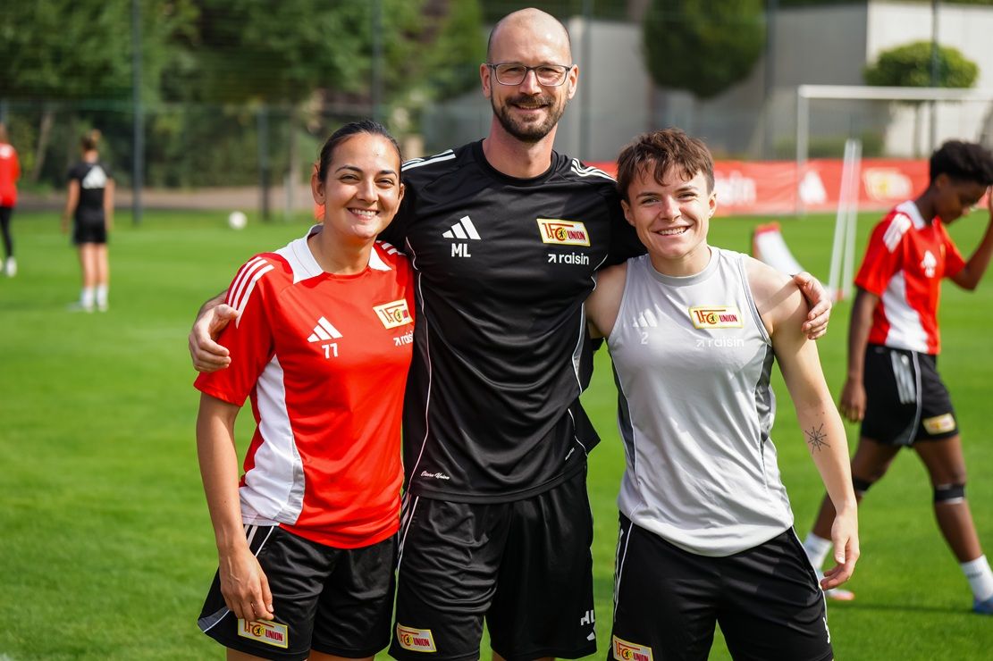 Drei Personen stehen auf einem Fußballfeld, zwei Spielerinnen in Trikots und ein Trainer in schwarzer Sportkleidung.