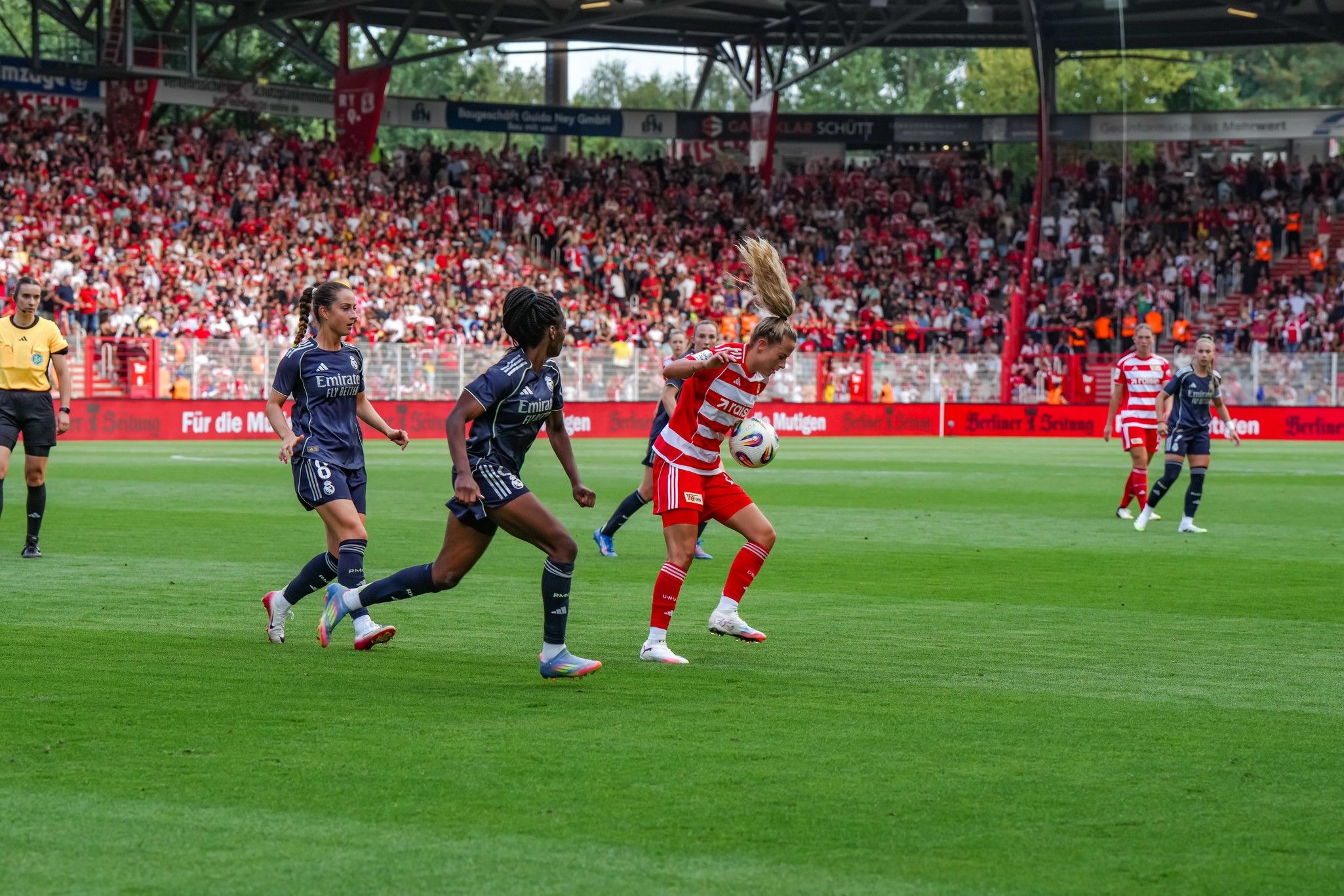 Spielszene aus einem Fußballspiel mit zwei Spielerinnen in blauem Trikot, die eine Spielerin in rot verfolgen, Hintergrund mit jubelnden Fans.