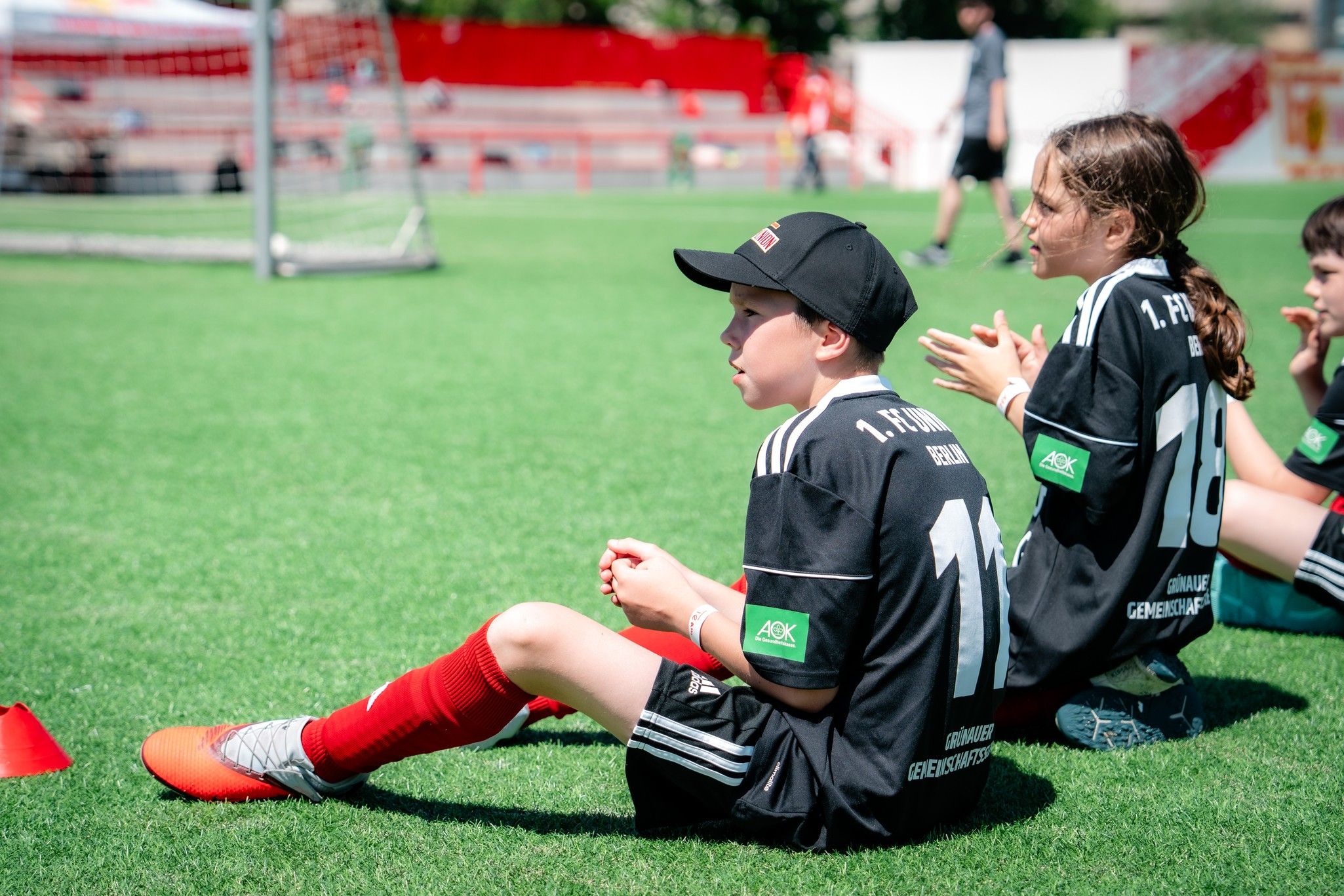 Zwei Kinder in Fußballtrikots sitzen auf einem Sportplatz, warten auf ihren Einsatz und schauen auf das Spielfeld.