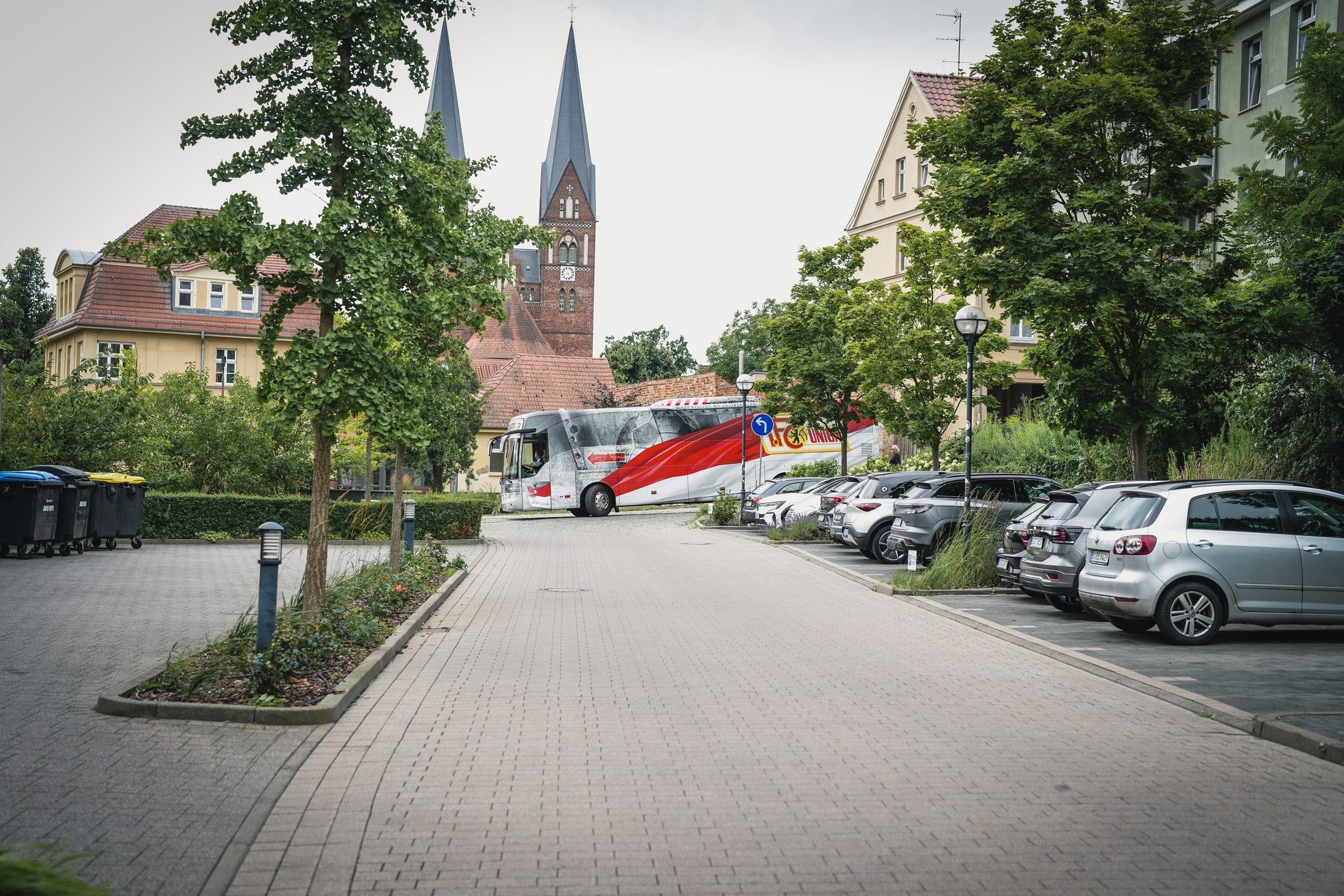 Ein Bus parkt in einer Straße mit Bäumen und geparkten Autos. Im Hintergrund sind Kirchenstützen zu sehen.