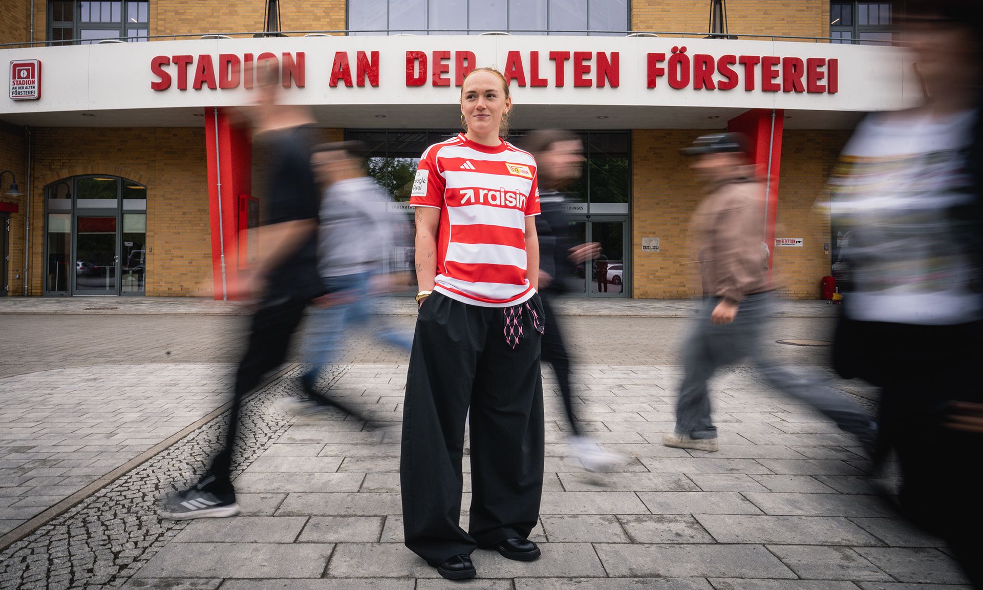Eine Person in einem rot-weißen Fußballtrikot steht vor dem Stadion An der Alten Försterei, während Menschen vorbeigehen.