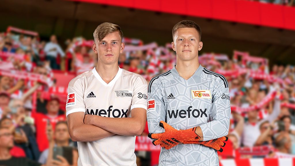 Zwei Fußballspieler im Stadion, einer in einem weißen Trikot, der andere in grauer Torwartausrüstung, mit Fans im Hintergrund.