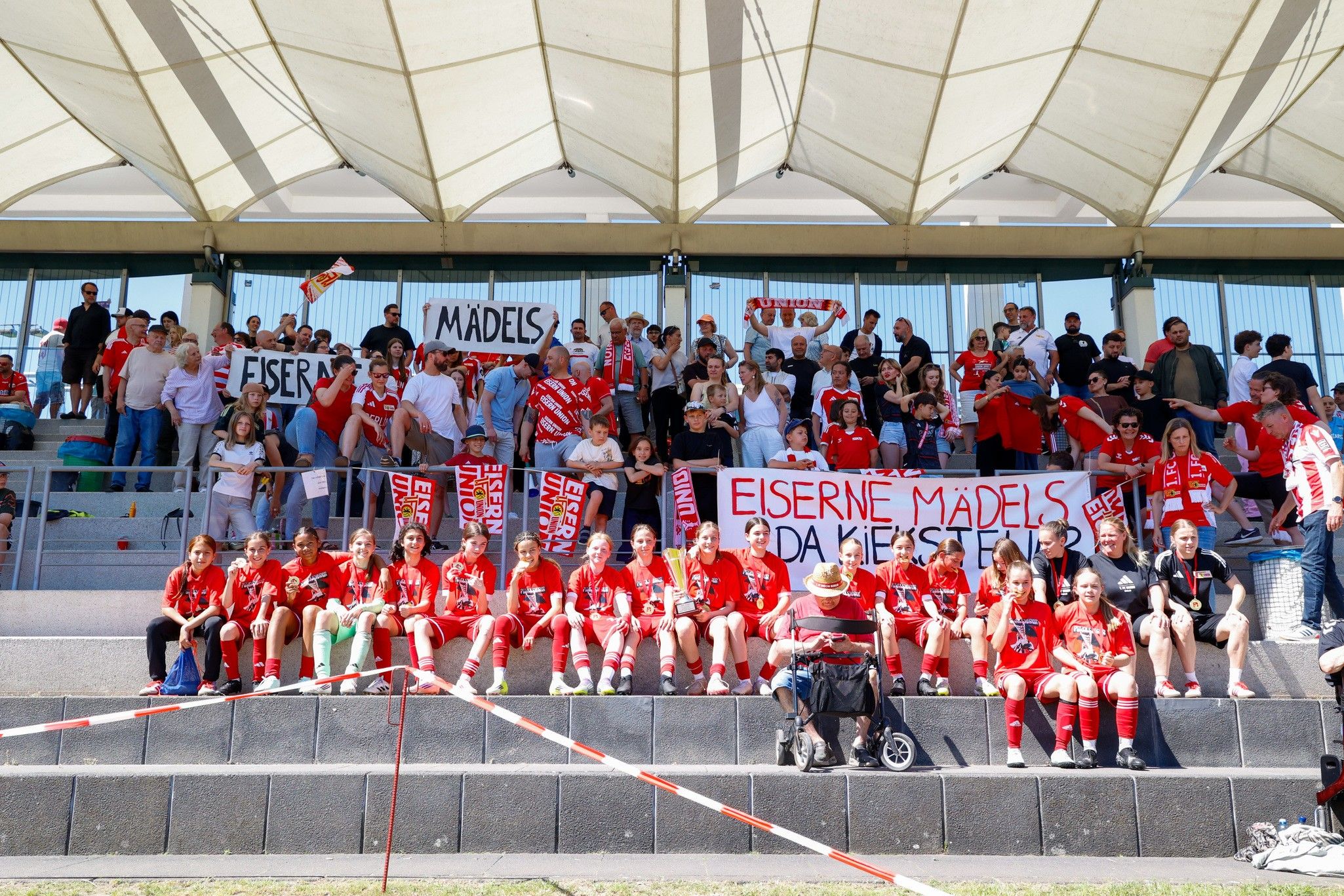 Fans einer Fußballmannschaft stehen und sitzen in einer Tribüne, während sie mit Schildern ihre Unterstützung zeigen.